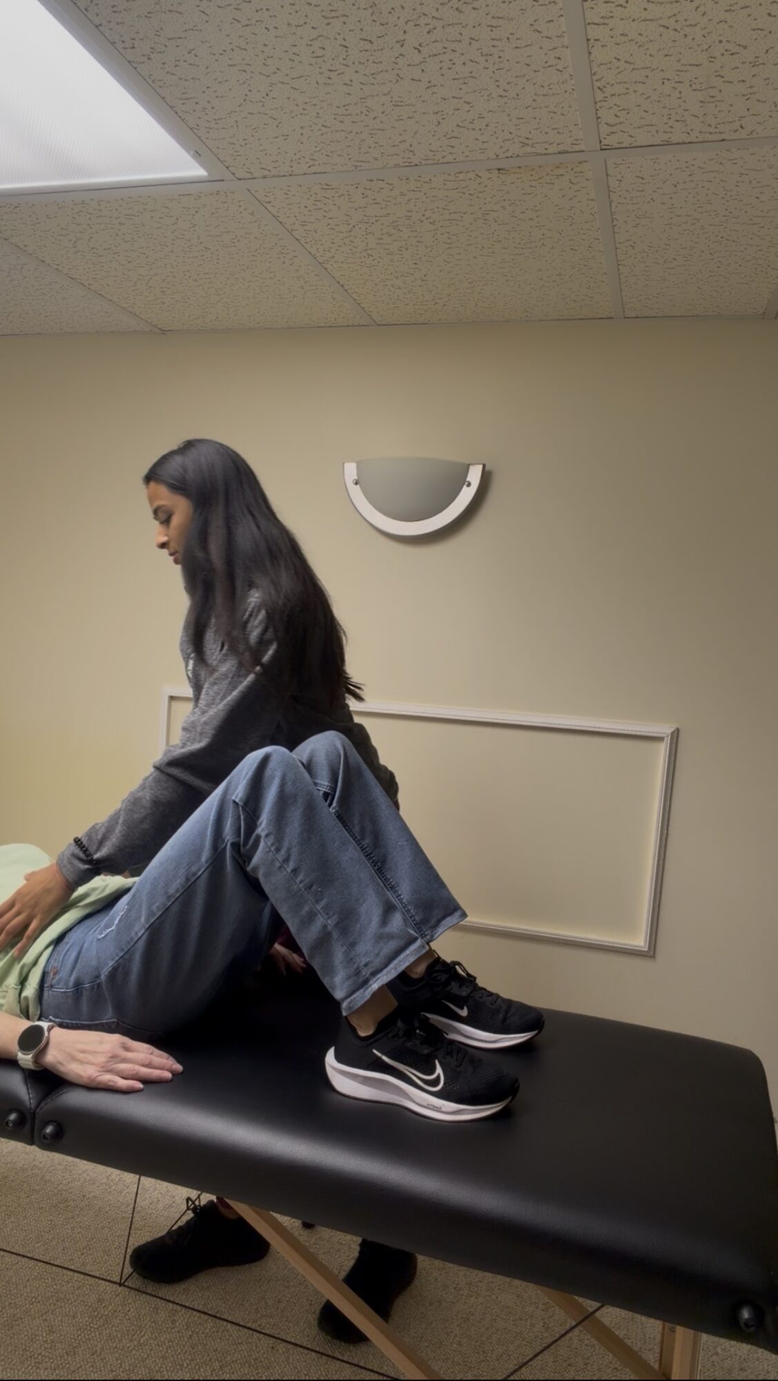 Person lying on a black examination table with legs crossed, in a room with beige walls and ceiling light.