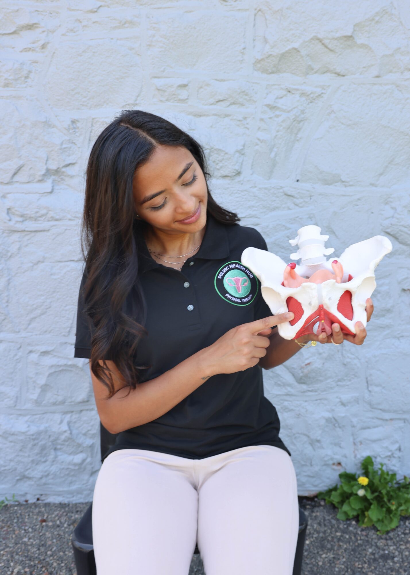 Woman with long dark hair holding a model of a human pelvis, pointing at it, standing outdoors against a white brick wall.