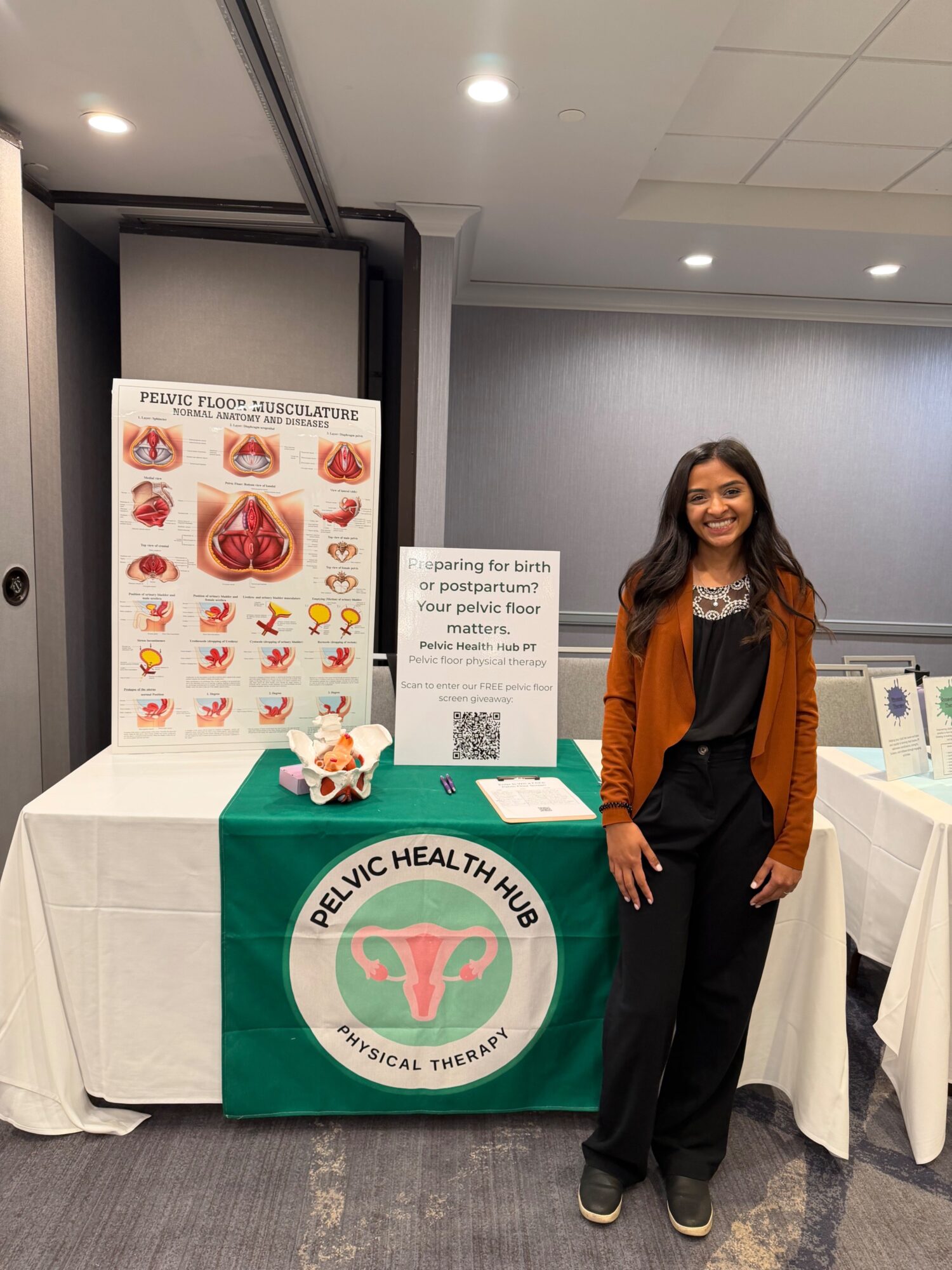 Woman standing next to a health information booth with a green tablecloth and a pelvic health logo, in an indoor conference setting.