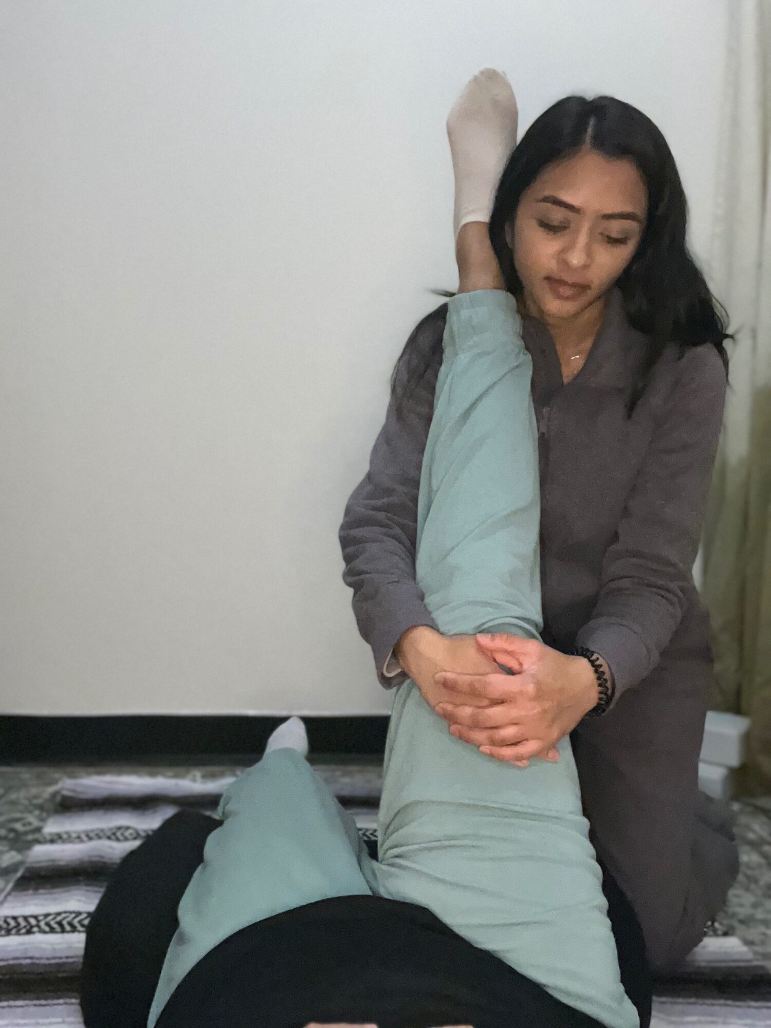 Woman with long dark hair gently holding a person's leg, which is raised and bent, in a room with a white wall.