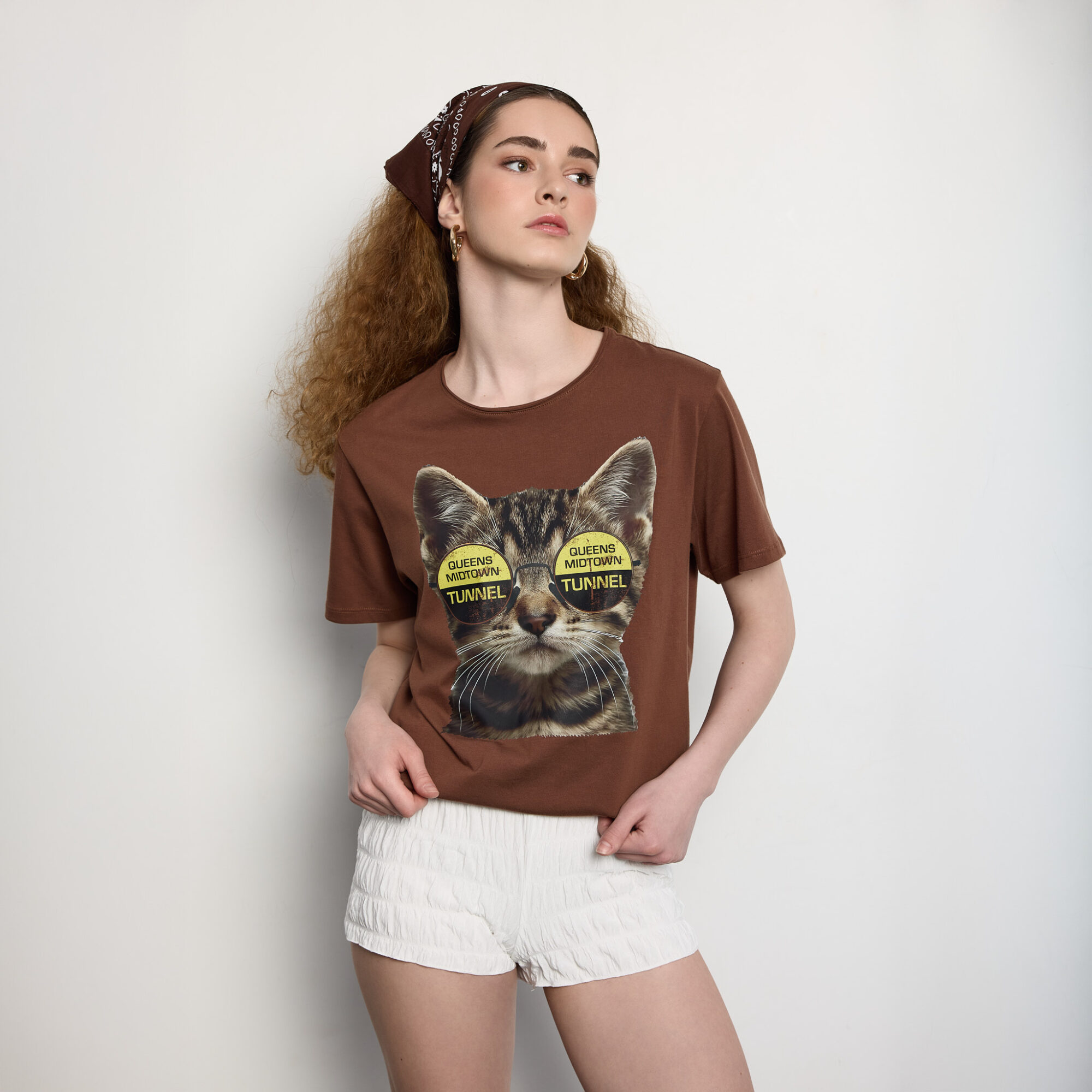 Young woman with curly hair wearing a brown T-shirt with a cat graphic and white shorts, standing against a white background.