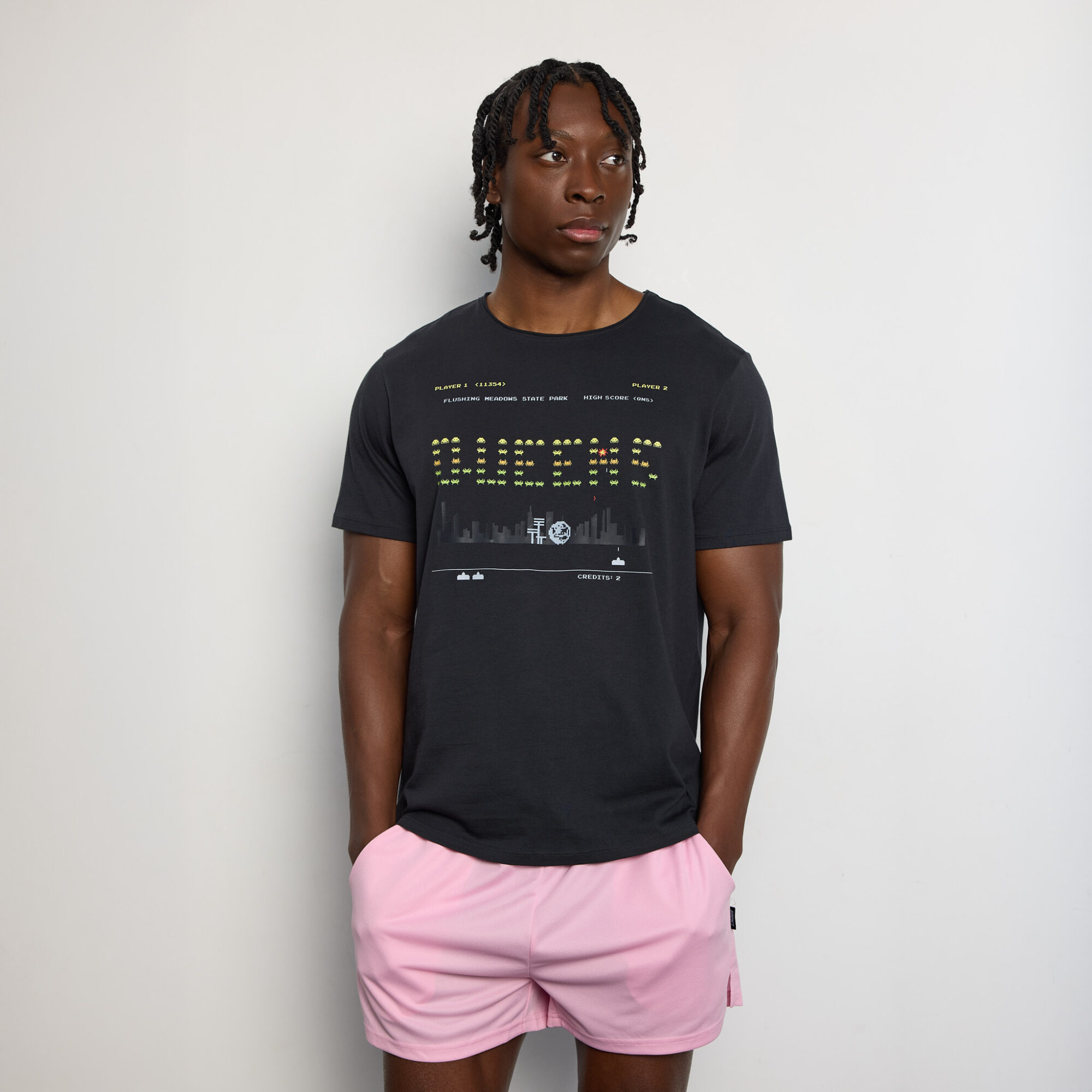 Person with short dreadlocks wearing a black T-shirt and pink shorts, standing against a plain white background.