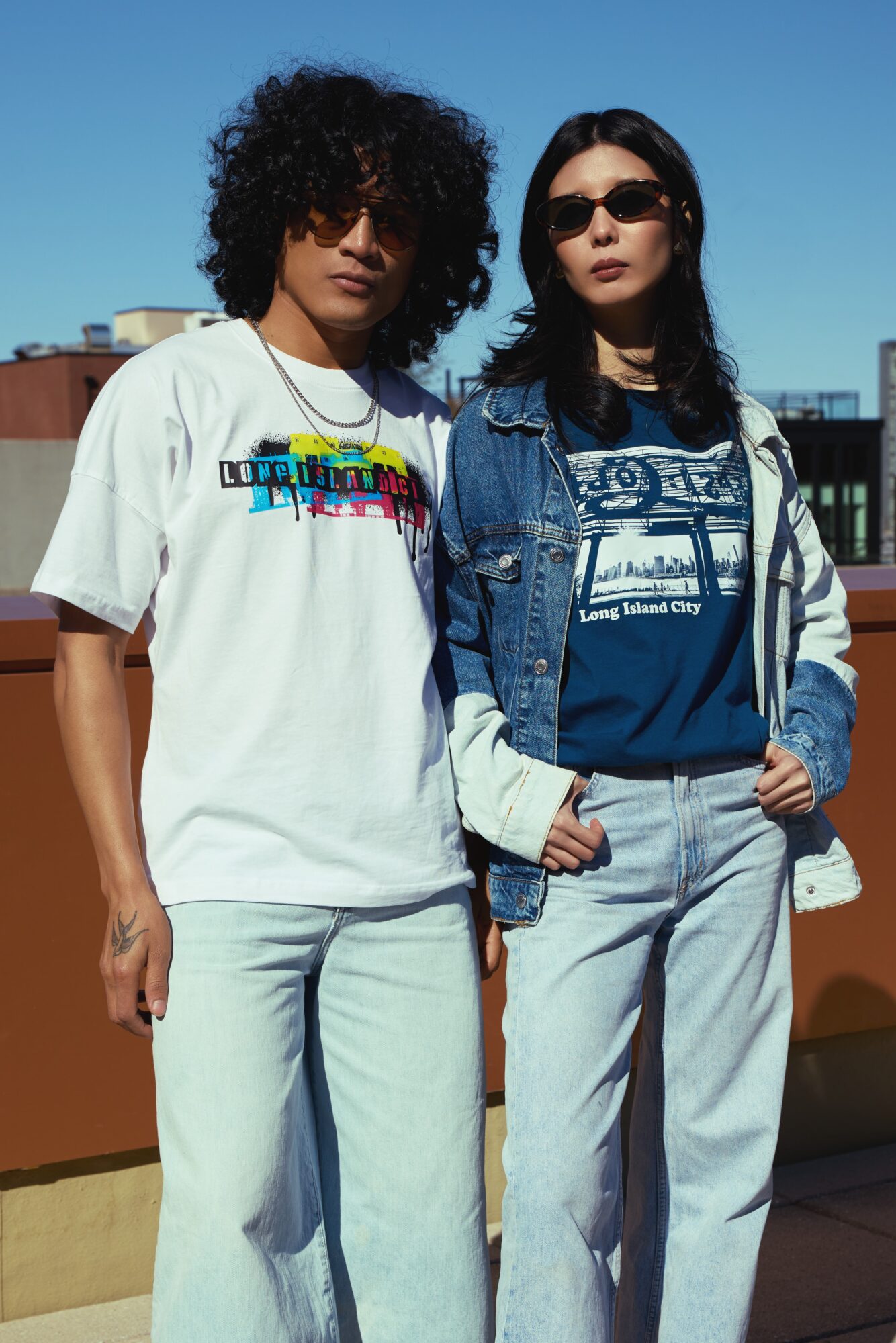 Two women standing outdoors against a cityscape background, one with curly hair and sunglasses, the other with straight hair and sunglasses, both in casual clothing.