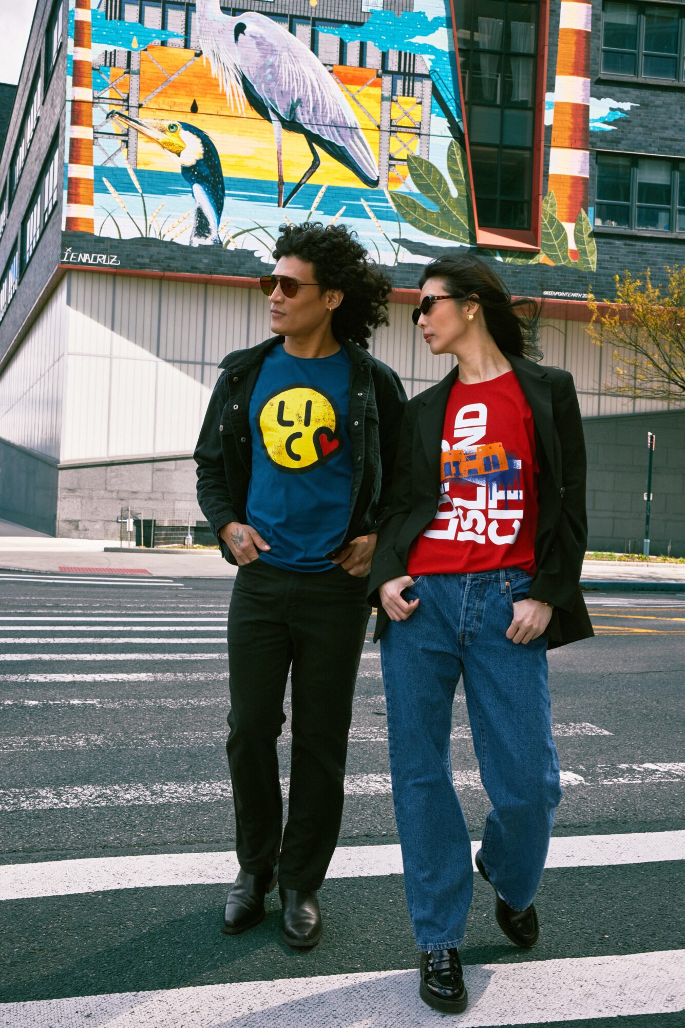 Two people walk across a crosswalk in an urban area with a colorful mural in the background.