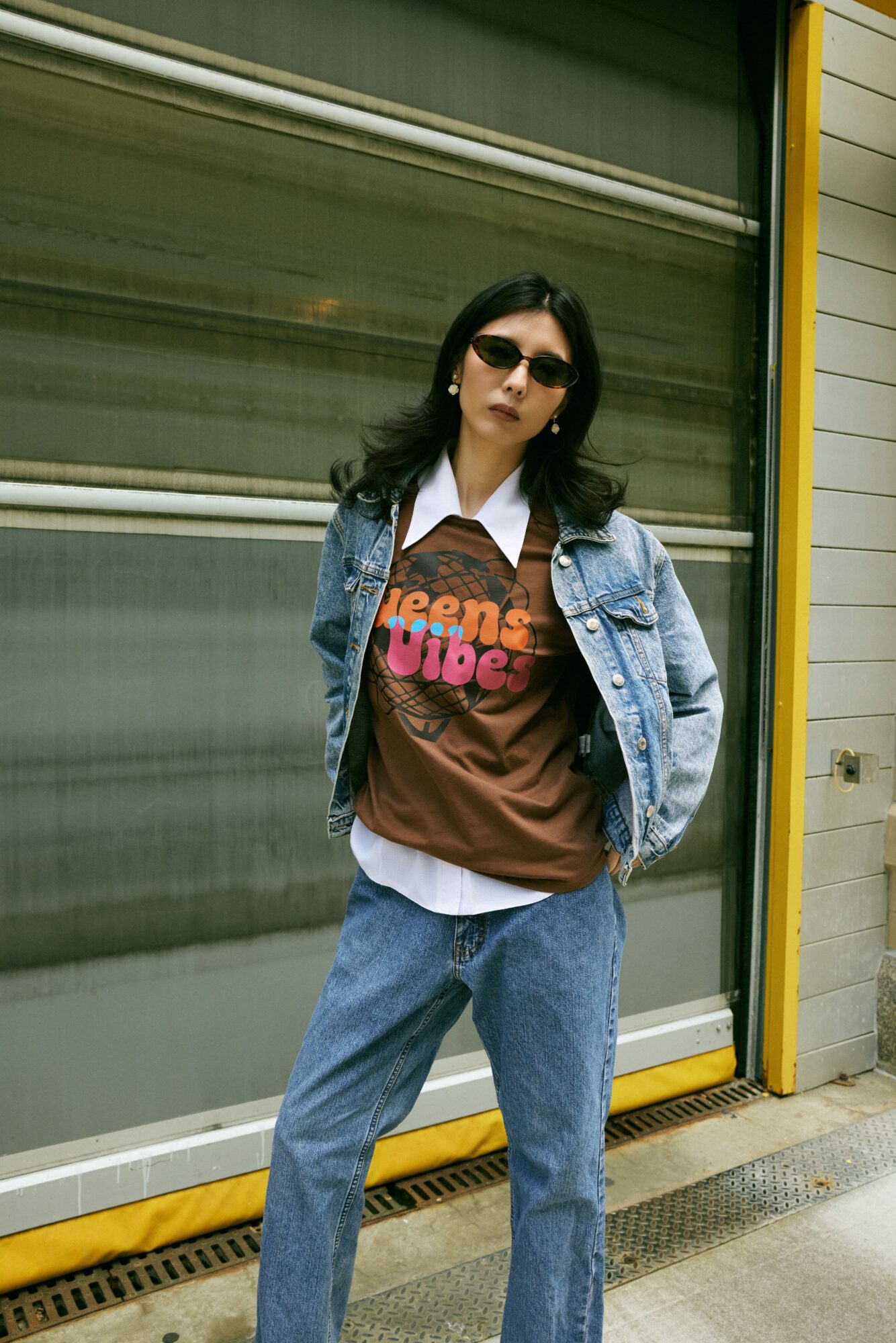 Woman with dark hair wearing sunglasses, denim jacket, brown graphic t-shirt, and jeans, standing outside near a yellow and gray building.