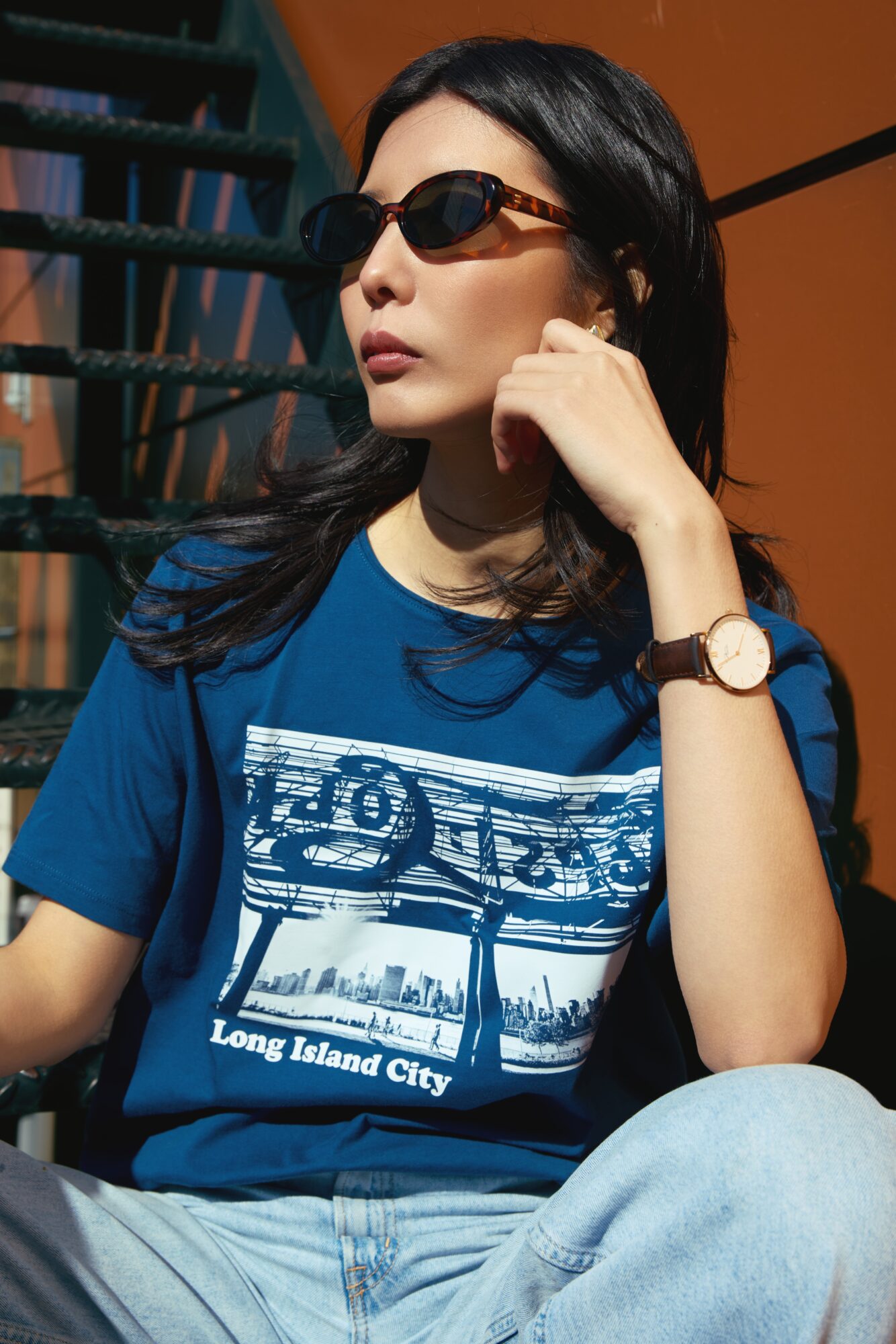 Woman with dark hair wearing sunglasses, a blue graphic T-shirt, and a watch, sitting outdoors.