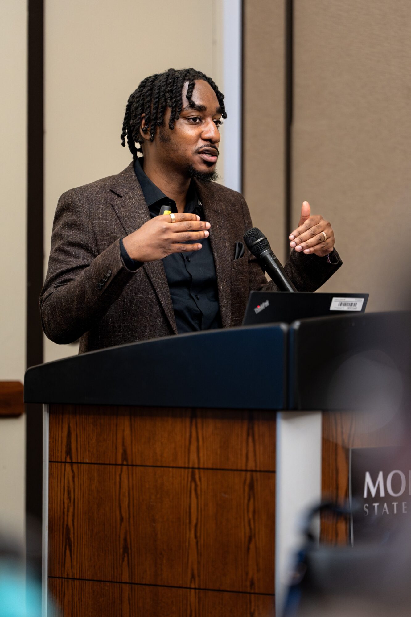 Person speaking at a podium, gesturing with hands, wearing a brown blazer and black shirt, in a room with beige walls.