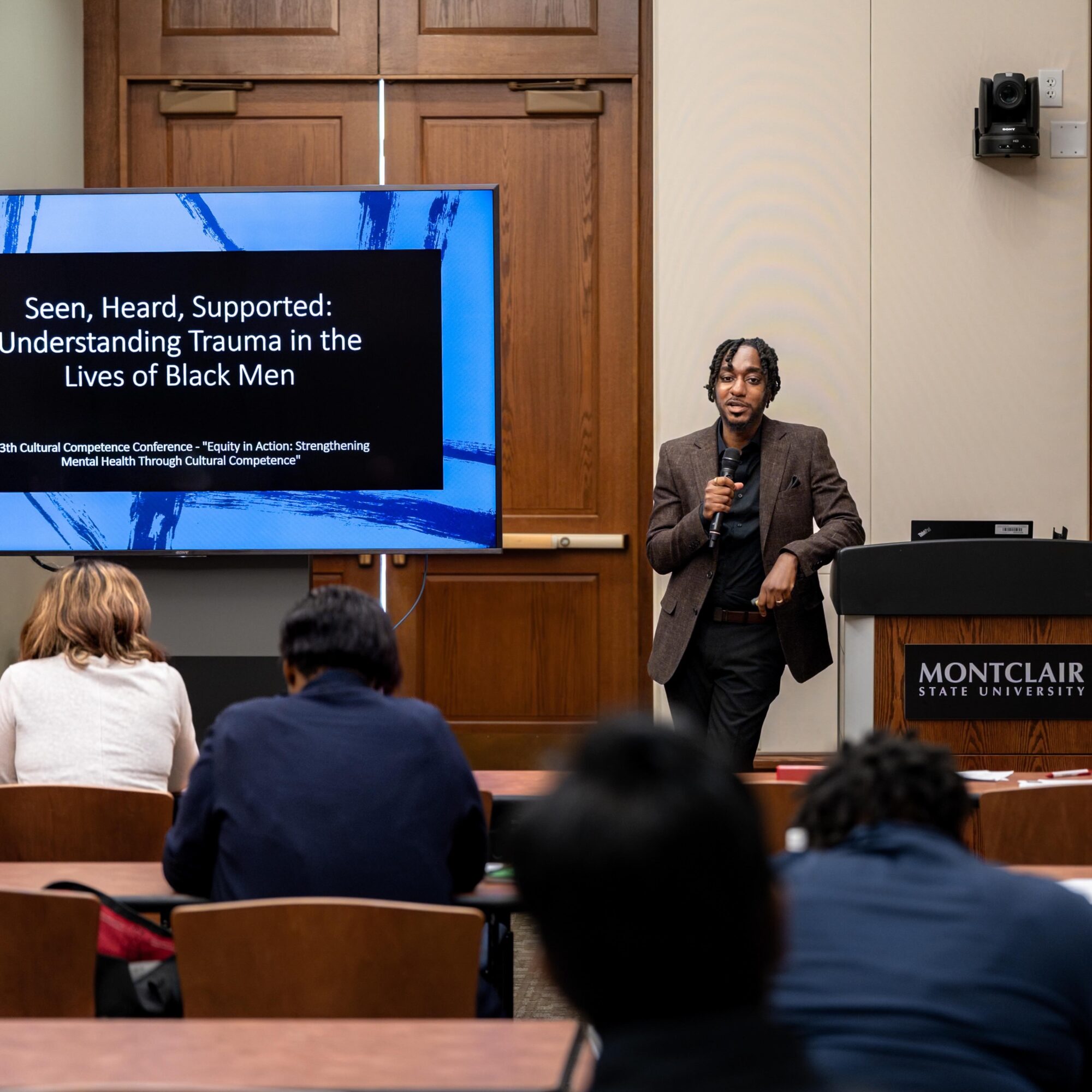 A woman stands at a podium in a classroom, giving a presentation to students seated facing her. A large screen displays a slide about trauma in Black men.