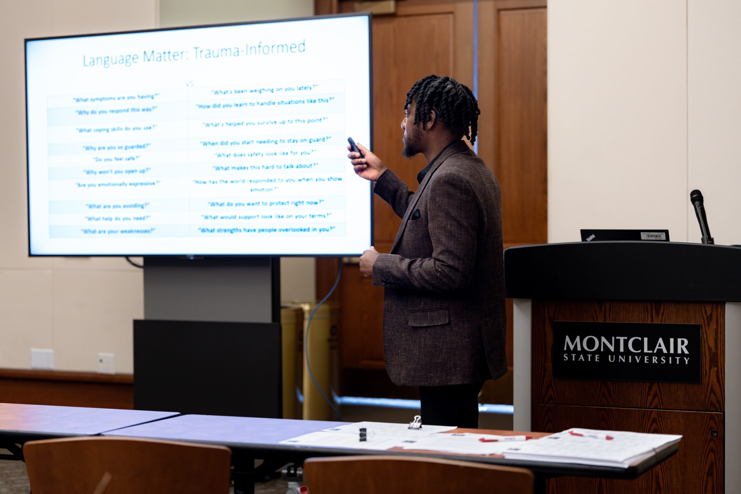Person presenting in front of a large screen with text, standing next to a podium at Montclair State University.