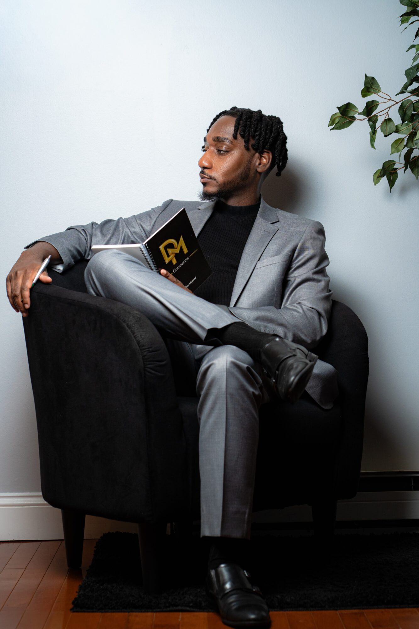 Man in gray suit reading a book while sitting in a black armchair, with a plant nearby.