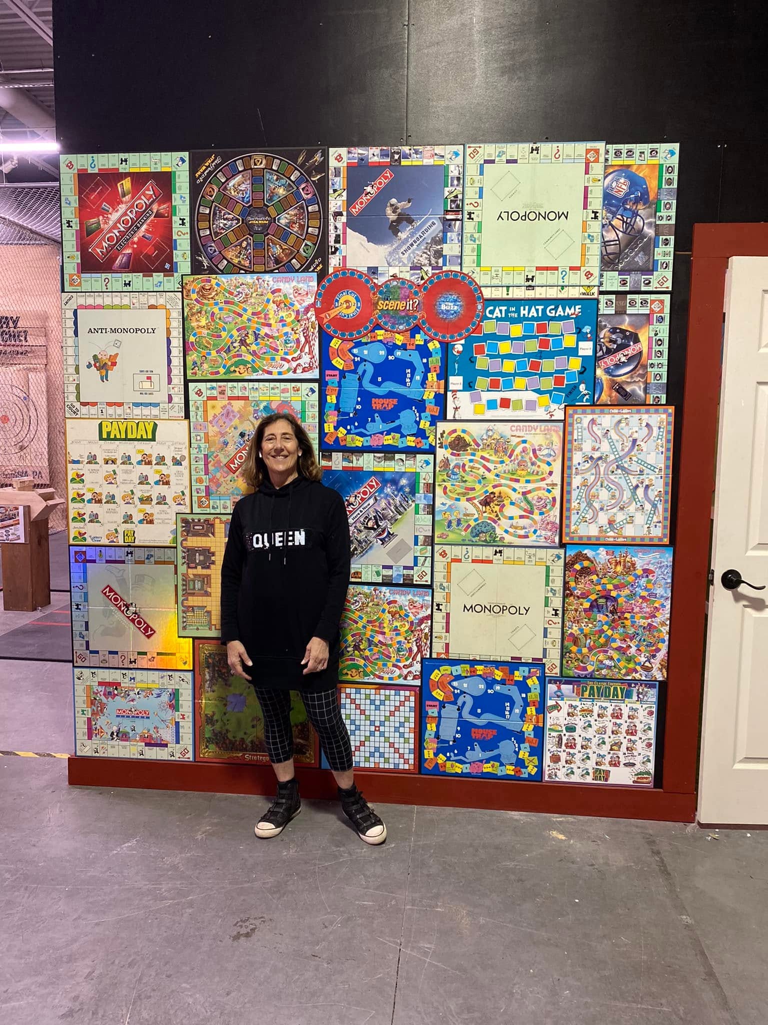 Person standing in front of a colorful display of board games on a wall, smiling, wearing a black hoodie and jeans.
