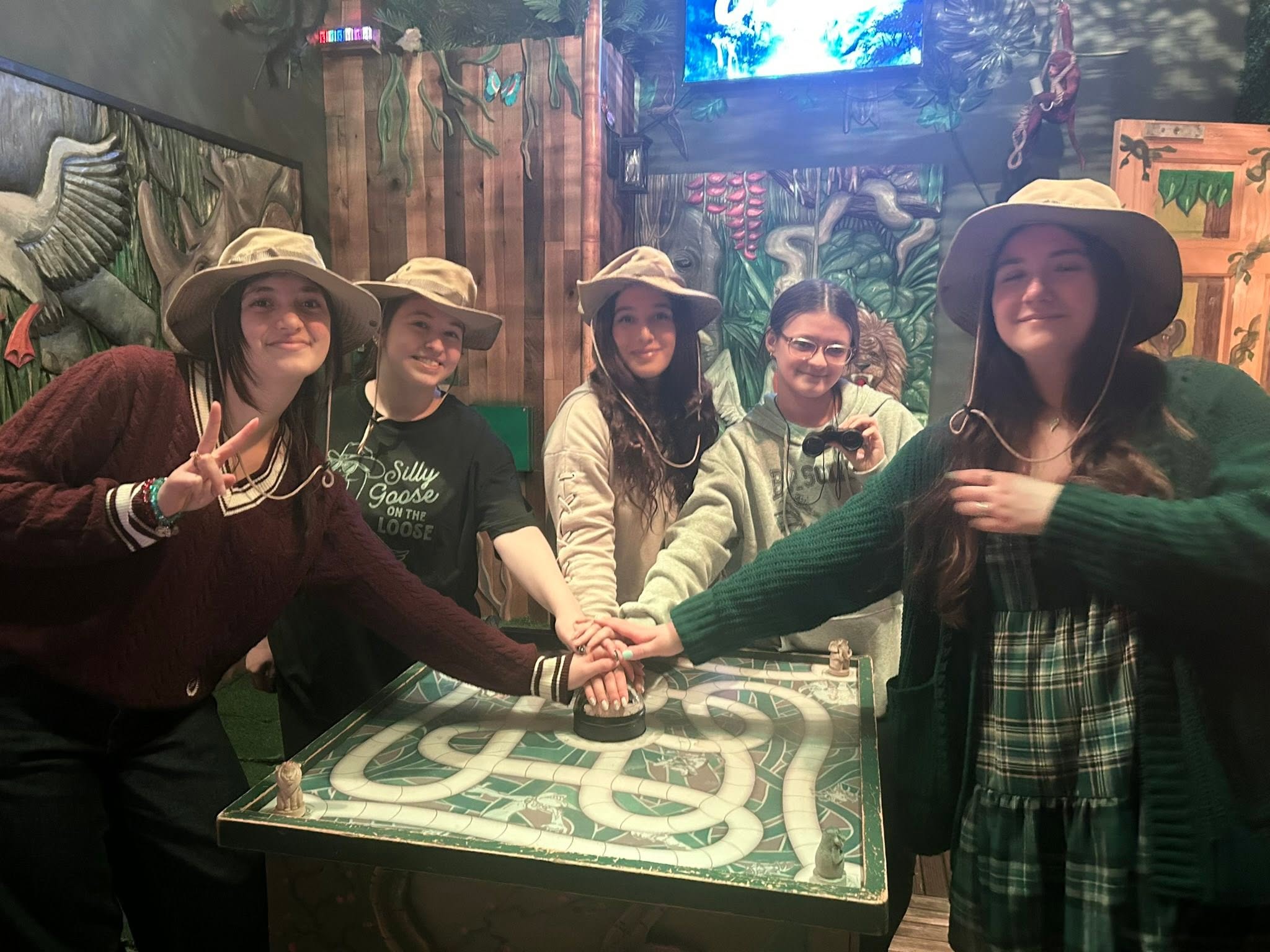 Five women wearing hats hold hands around a maze table in a decorated indoor setting.