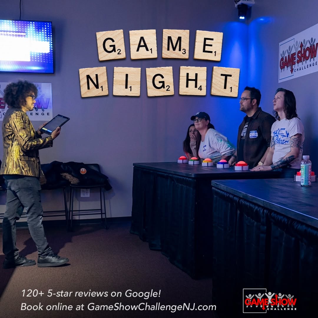Four people stand behind a table with game show buttons, a woman with a tablet, and Scrabble tiles spelling 'GAME NIGHT' on the wall.