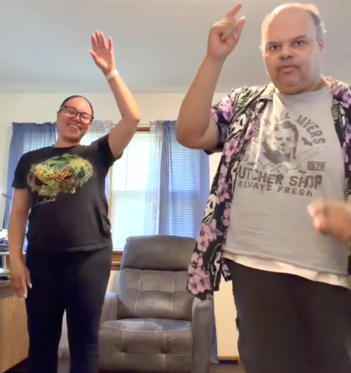 Two people standing indoors, raising their hands, smiling, with a window and curtains in the background.