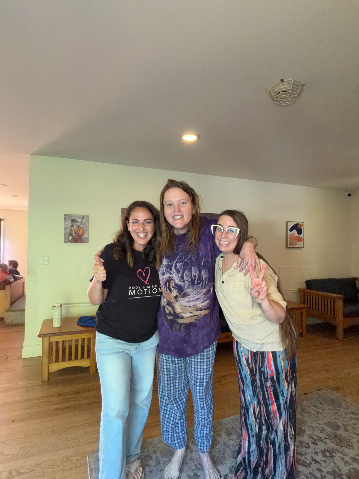 Three smiling women standing together indoors, arms around each other, in a room with light green walls and wooden furniture.