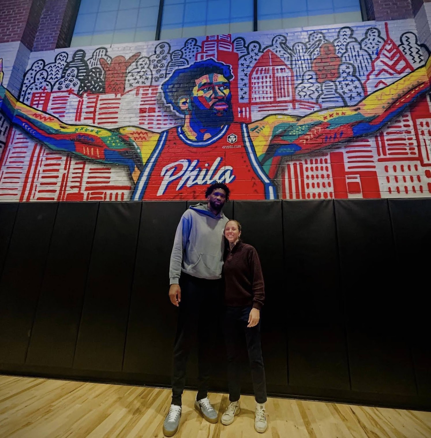 Two people stand in front of a colorful mural of a man with outstretched arms, with cityscape background, in an indoor setting.