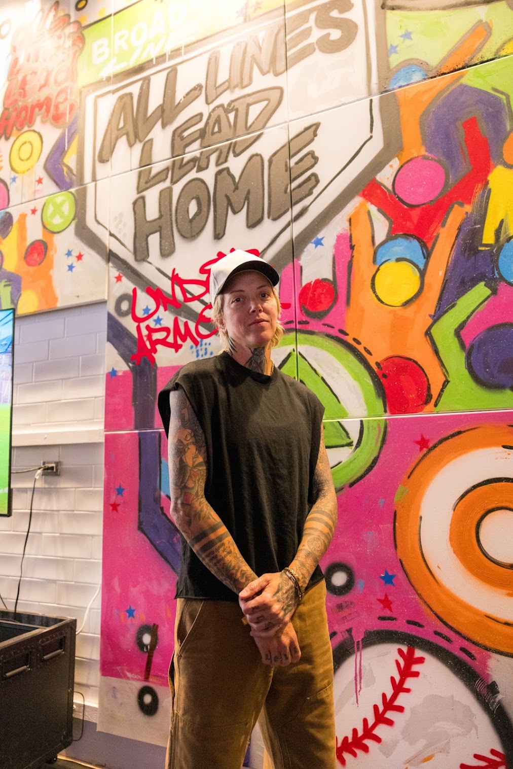 Person with tattoos wearing a black shirt and cap standing in front of colorful graffiti wall with text 'Allium's Lead Home'.