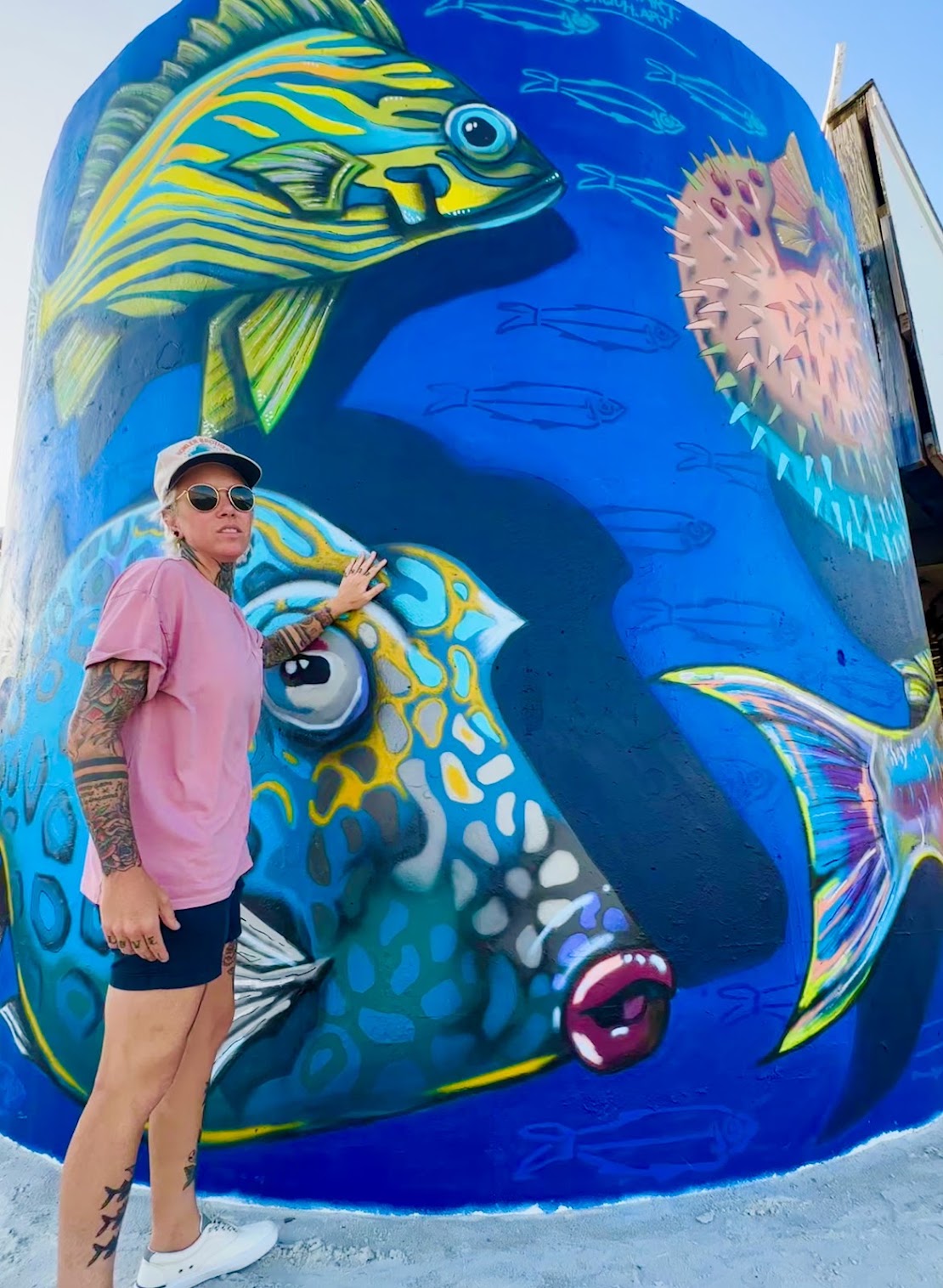 Person wearing sunglasses and pink shirt standing next to a colorful mural of fish and sea creatures on a wall.