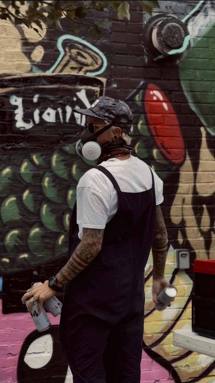 Person with tattoos and headphones painting a colorful mural on a wall, holding a spray can in one hand.