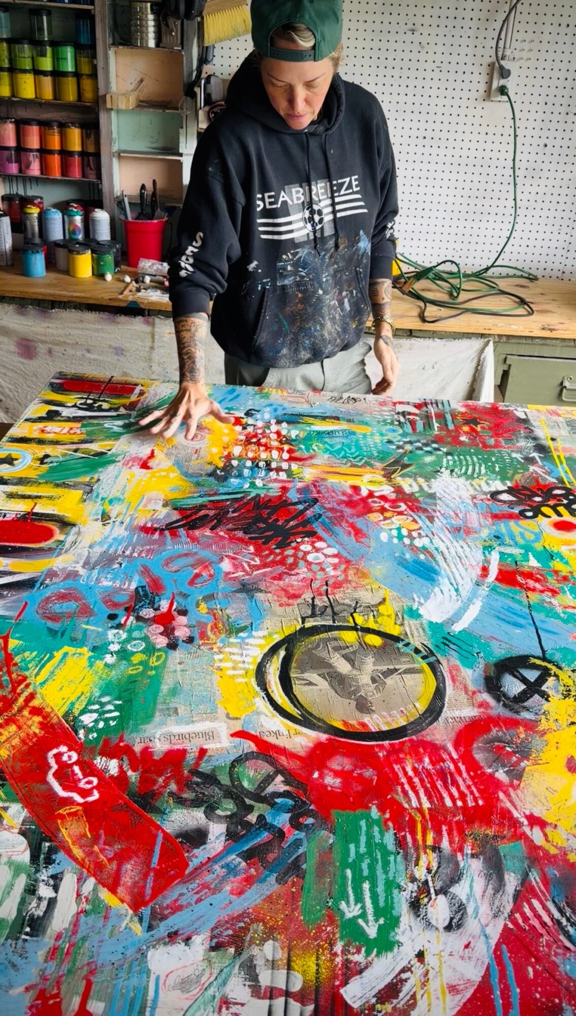 Person working on colorful abstract painting in studio with shelves and tools nearby.
