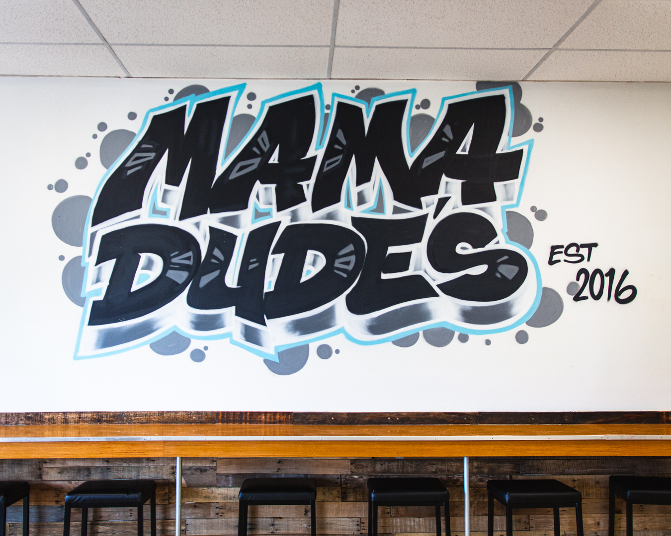 Graffiti-style text reads 'Mama Dudes' with 'EST 2016' on a white wall, above a wooden bar with stools.