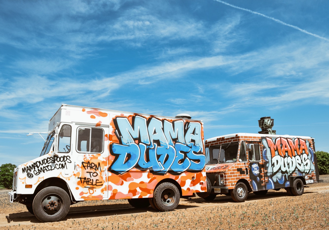 Two trucks with graffiti art reading 'Mama Dudes' parked on grass under a blue sky.