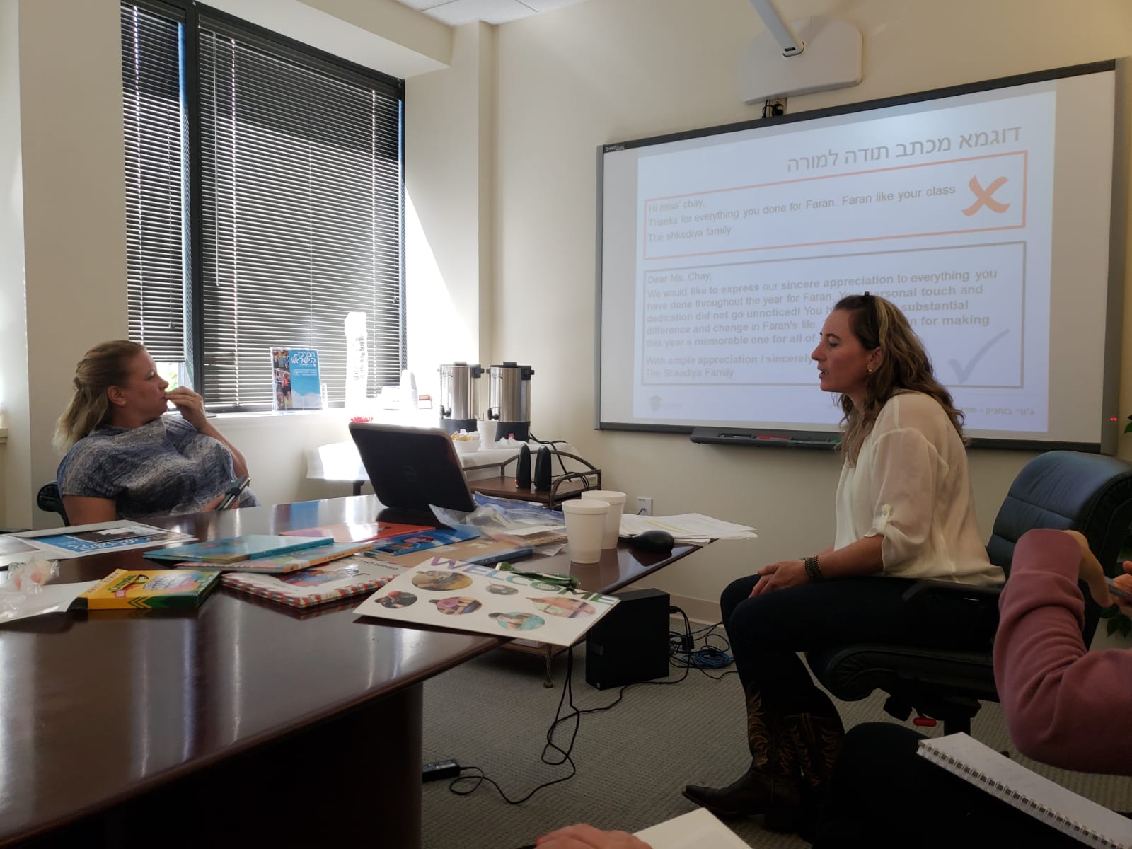 Two women in a meeting room with a large screen displaying text, a window with blinds, and a table with papers and a laptop.