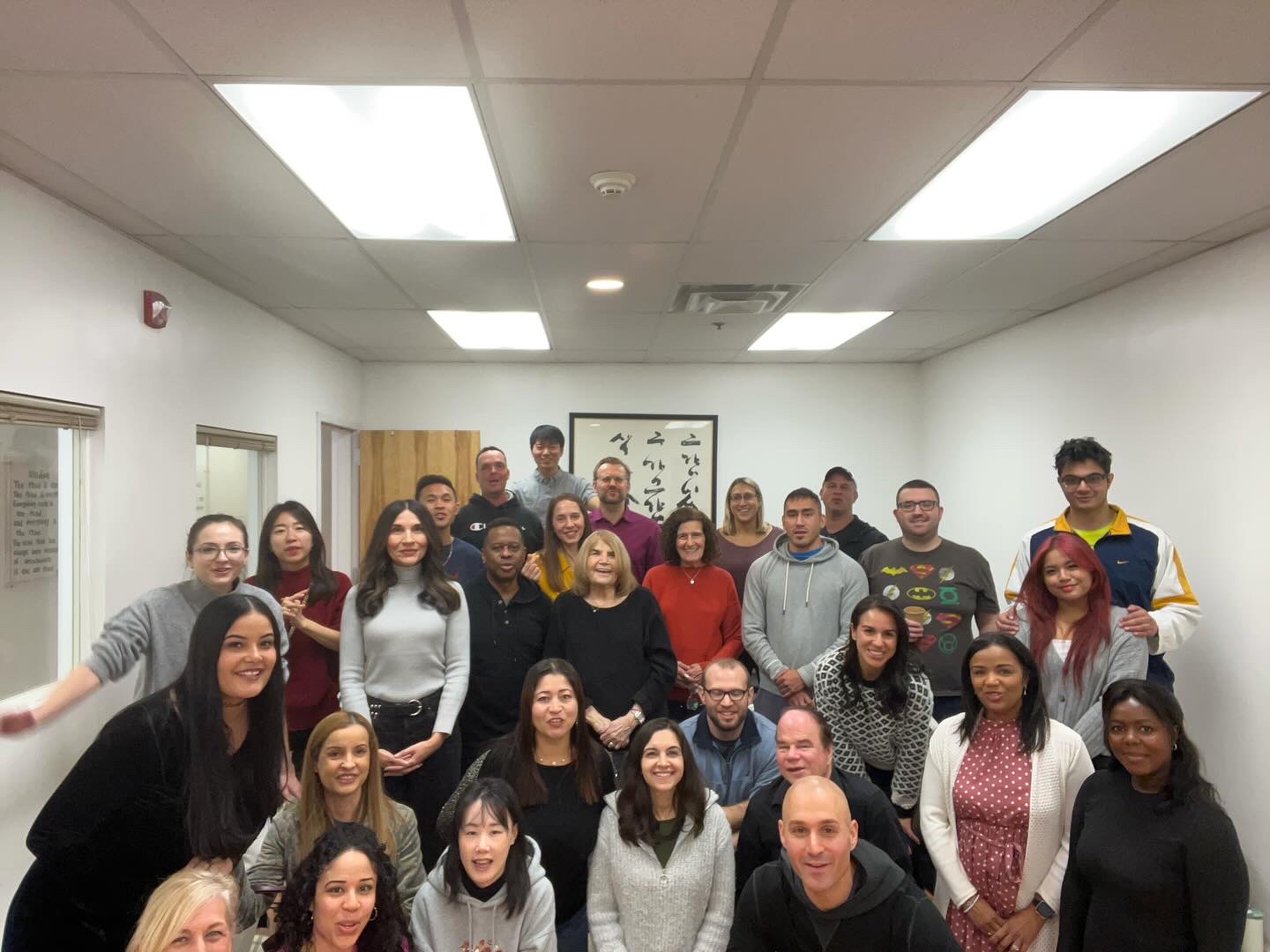 Group of 24 people standing in a classroom, smiling, with white walls and ceiling lights.