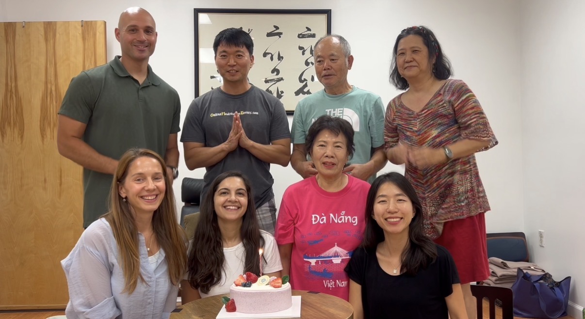 Group of seven people gathered around a table with a cake, smiling indoors, with a framed calligraphy artwork on the wall.