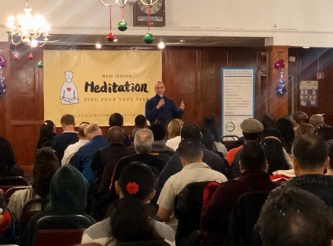 Person speaking on stage in front of a seated audience, with a banner reading 'Meditation' behind them.