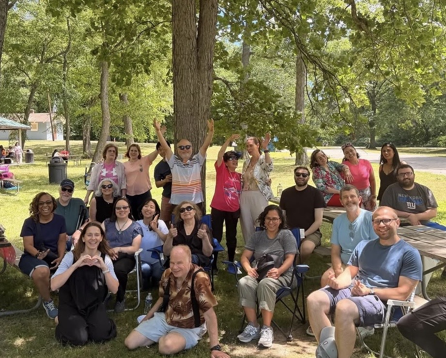 Group of people outdoors sitting and standing under trees, some making heart shapes with hands, in a park setting.