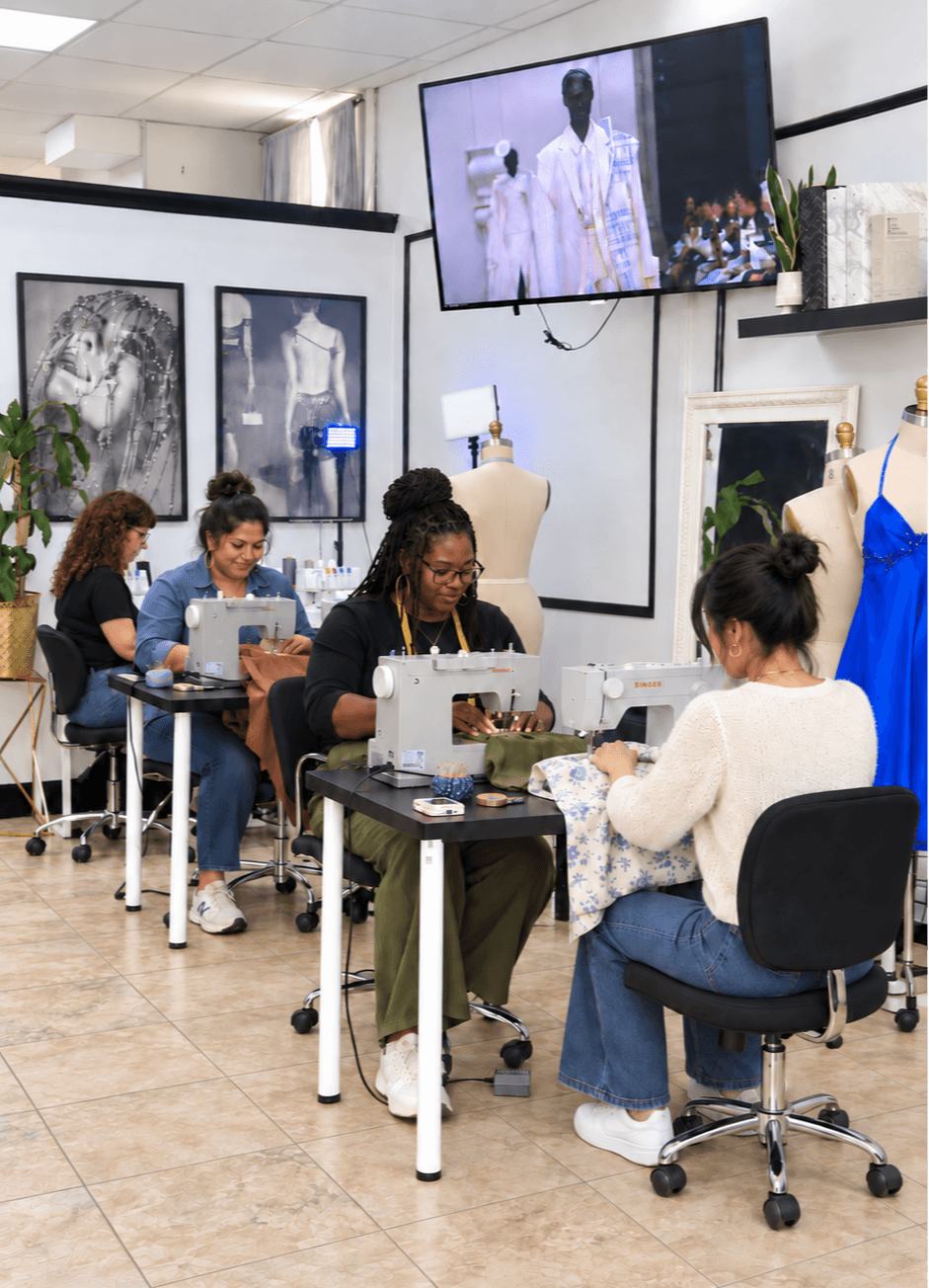 People sit at tables working on sewing projects in a room with clothing displays and artwork, a TV shows a person in white.
