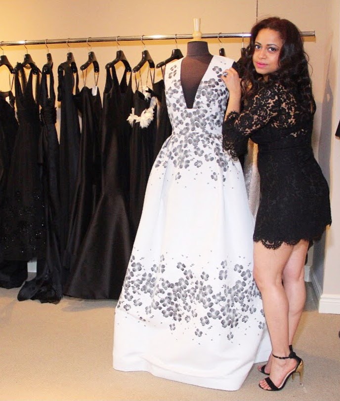 Woman in black lace dress standing next to a dress form with a white gown decorated with black floral patterns, black dresses hanging behind.