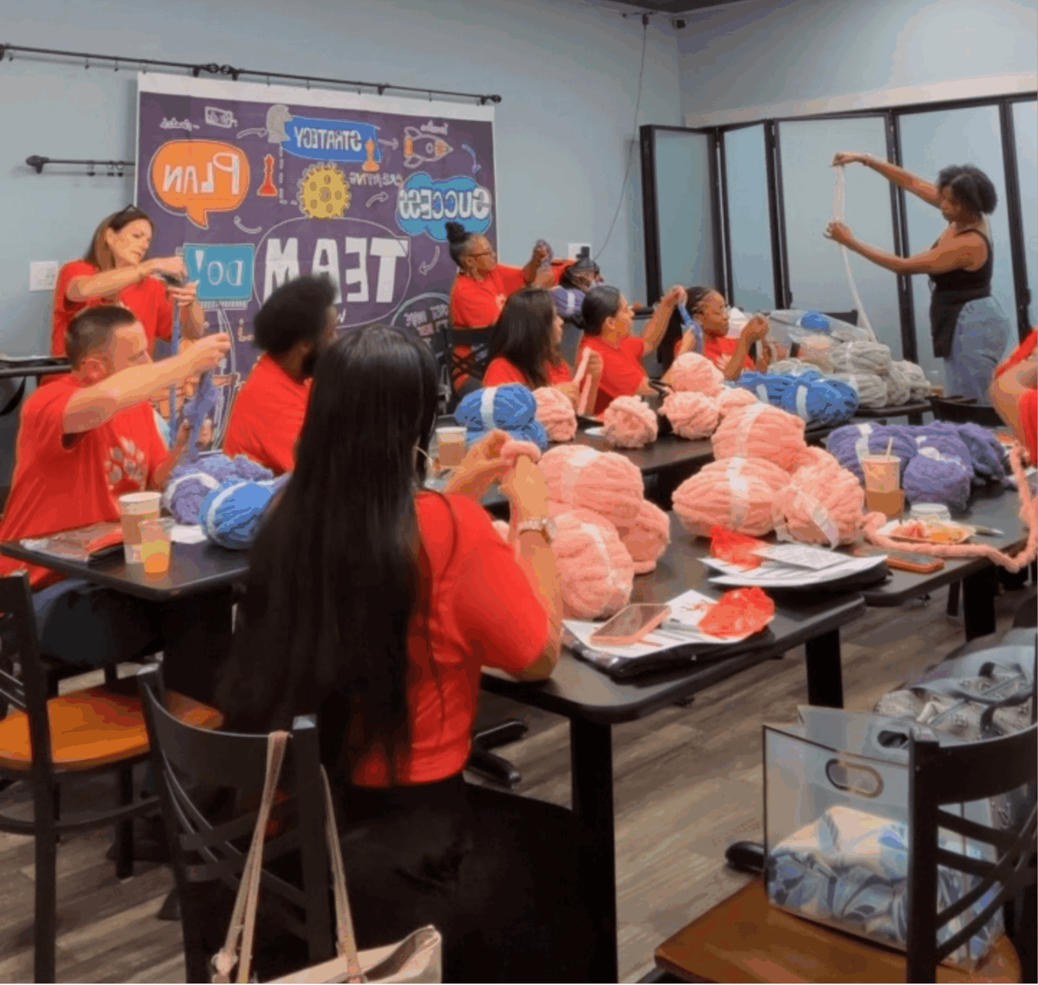 Group of people in a classroom participating in a knitting workshop, with yarn and knitting supplies on tables.