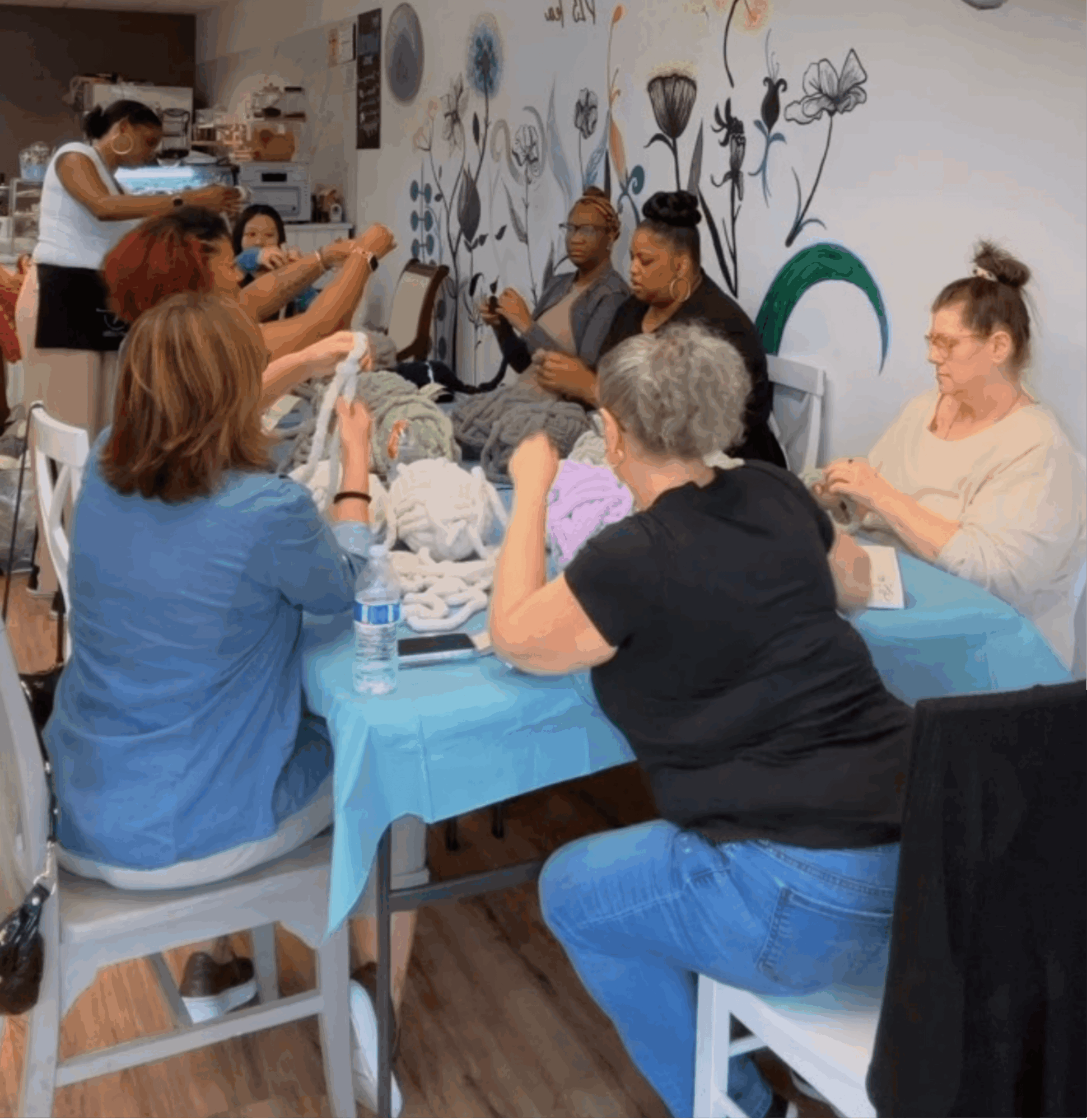 Group of women sitting around a table, some using phones, in a room with floral wall art.