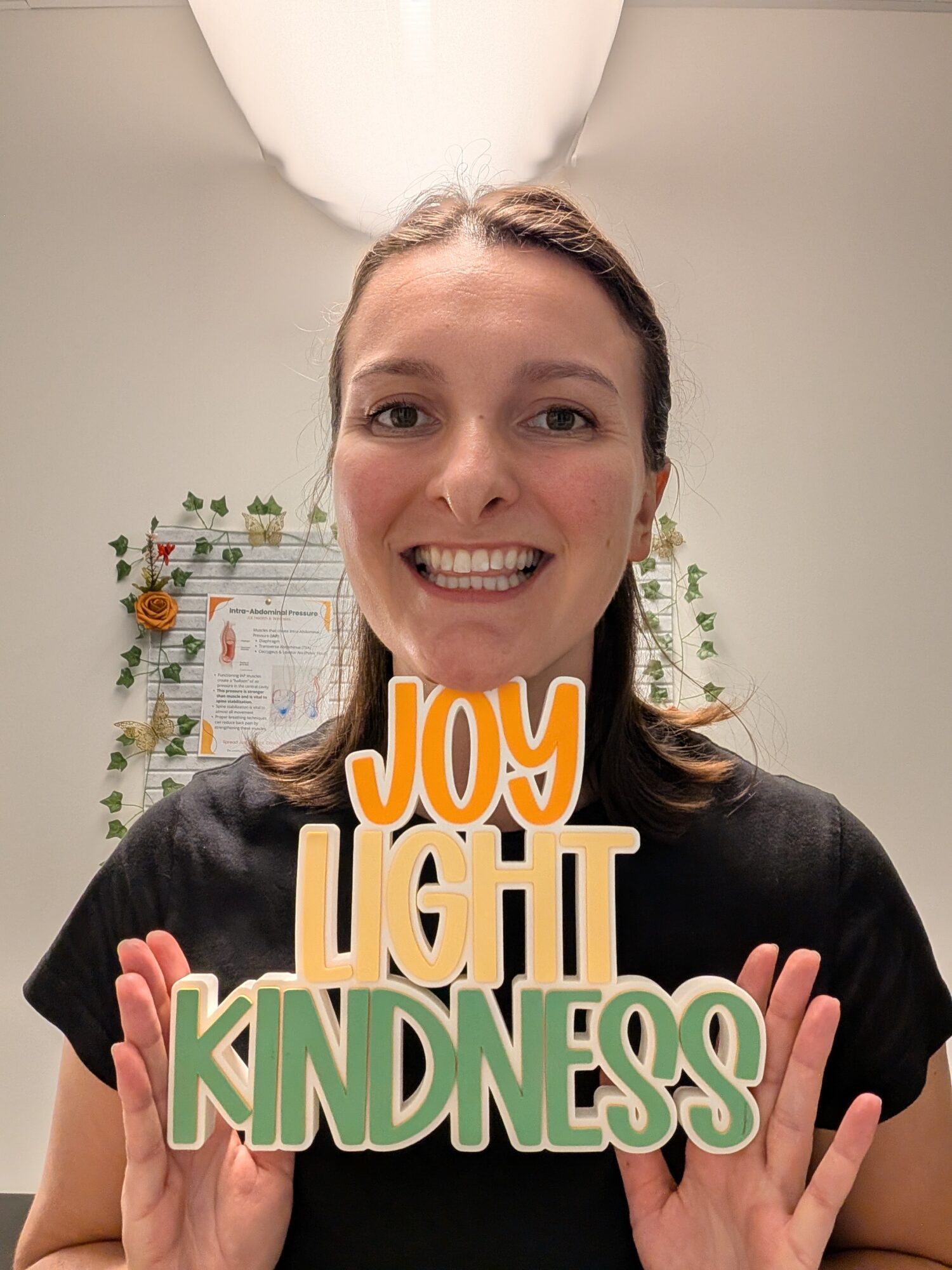 Smiling woman holding signs that say 'Joy,' 'Light,' and 'Kindness' in front of a decorated wall.