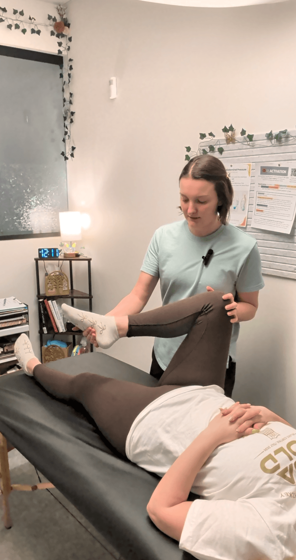 Person lying on a medical examination table with legs raised, another person standing nearby in a room with books and plants.