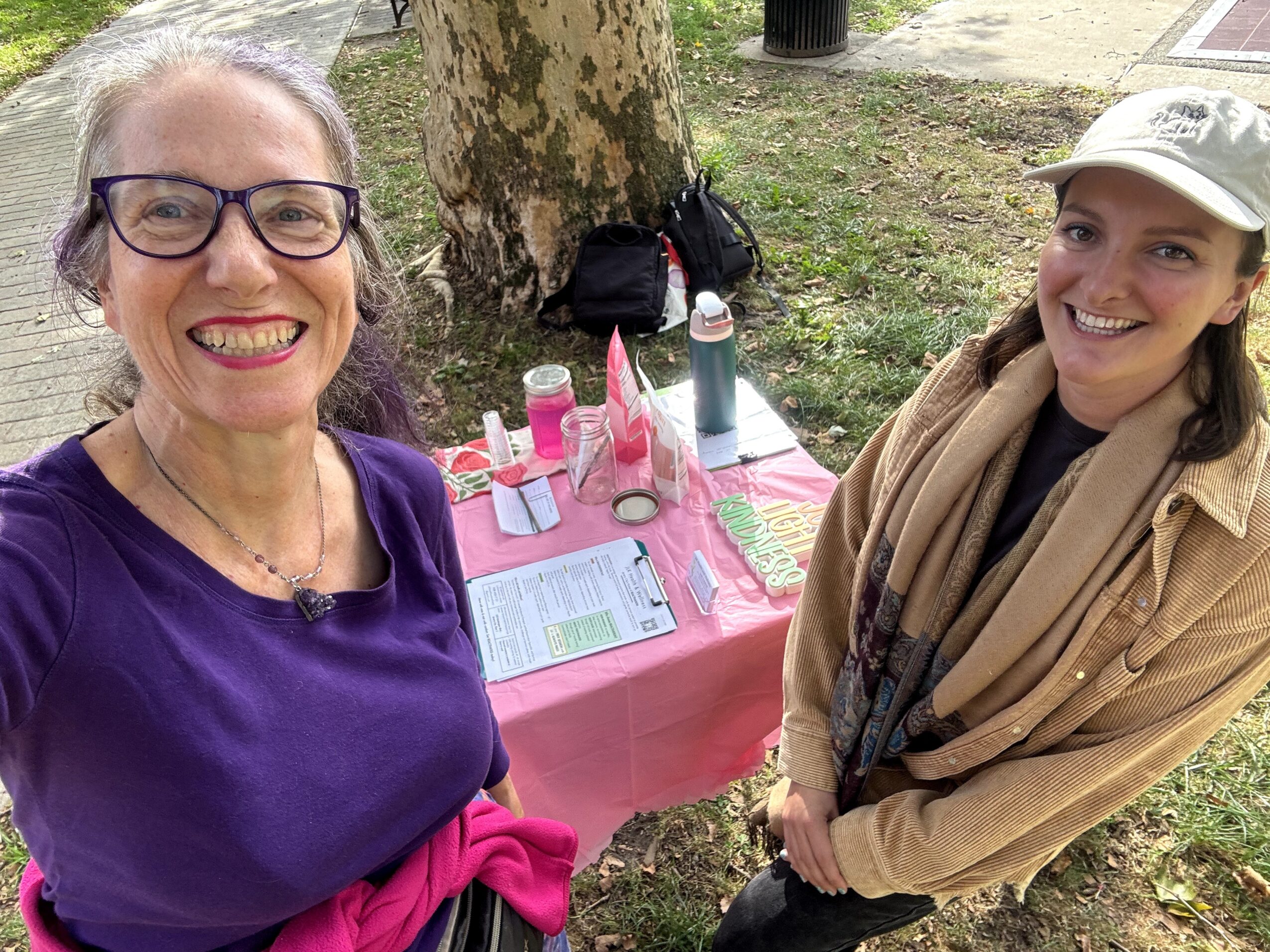Two women smiling outdoors near a pink table with drinks and papers, one wearing glasses and a purple top, the other in a beige coat and cap.