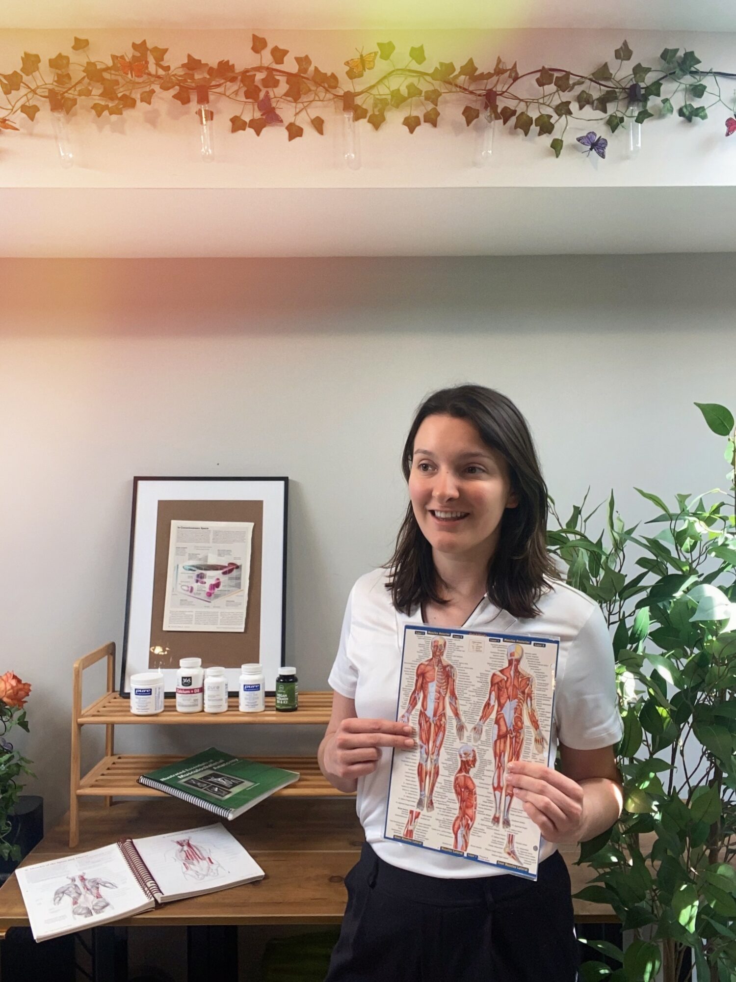 Young woman holding a diagram of human muscles, standing in a room with plants and a wooden shelf, smiling.