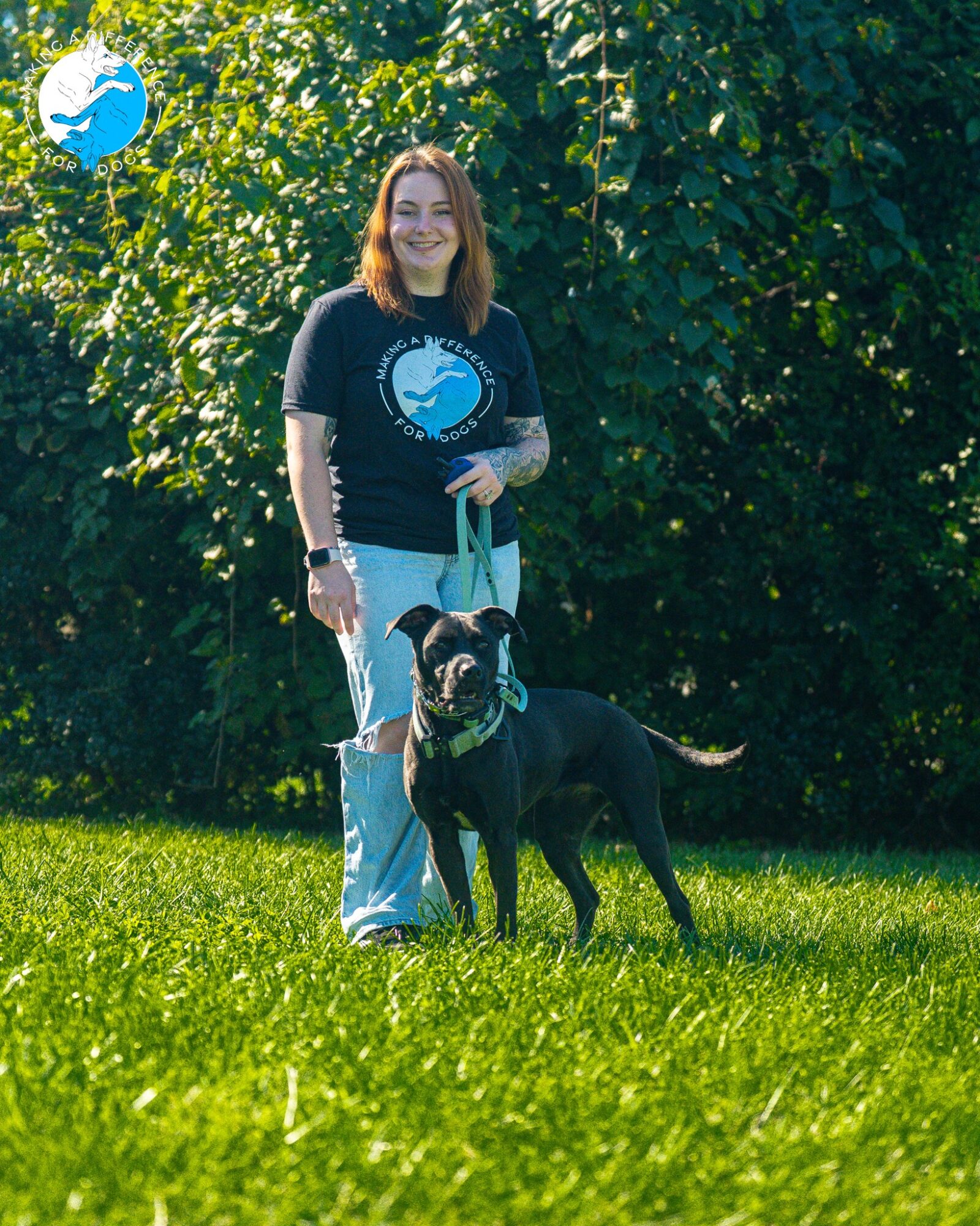 Woman standing outdoors with a black dog on a leash, green trees in background, smiling, sunny day.