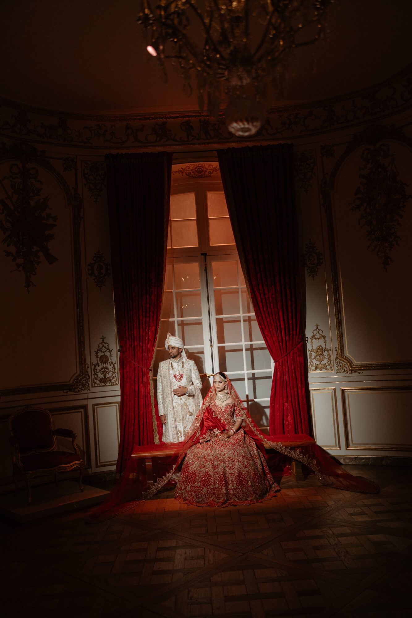Three people in elaborate historical costumes pose in front of a large window with red curtains, inside a decorated room.