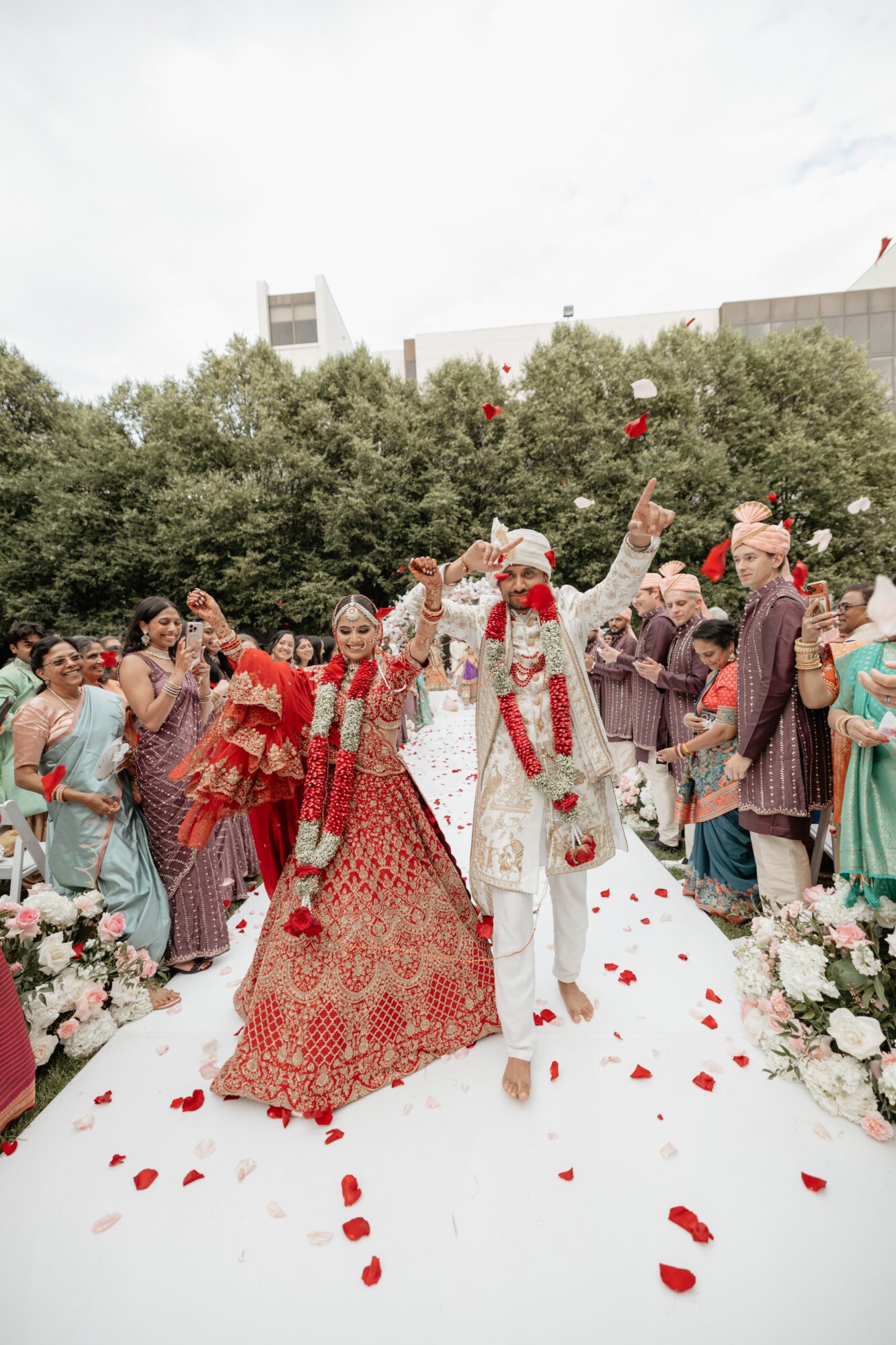 A bride and groom walk down an outdoor aisle surrounded by guests, with rose petals on the ground and trees in the background.