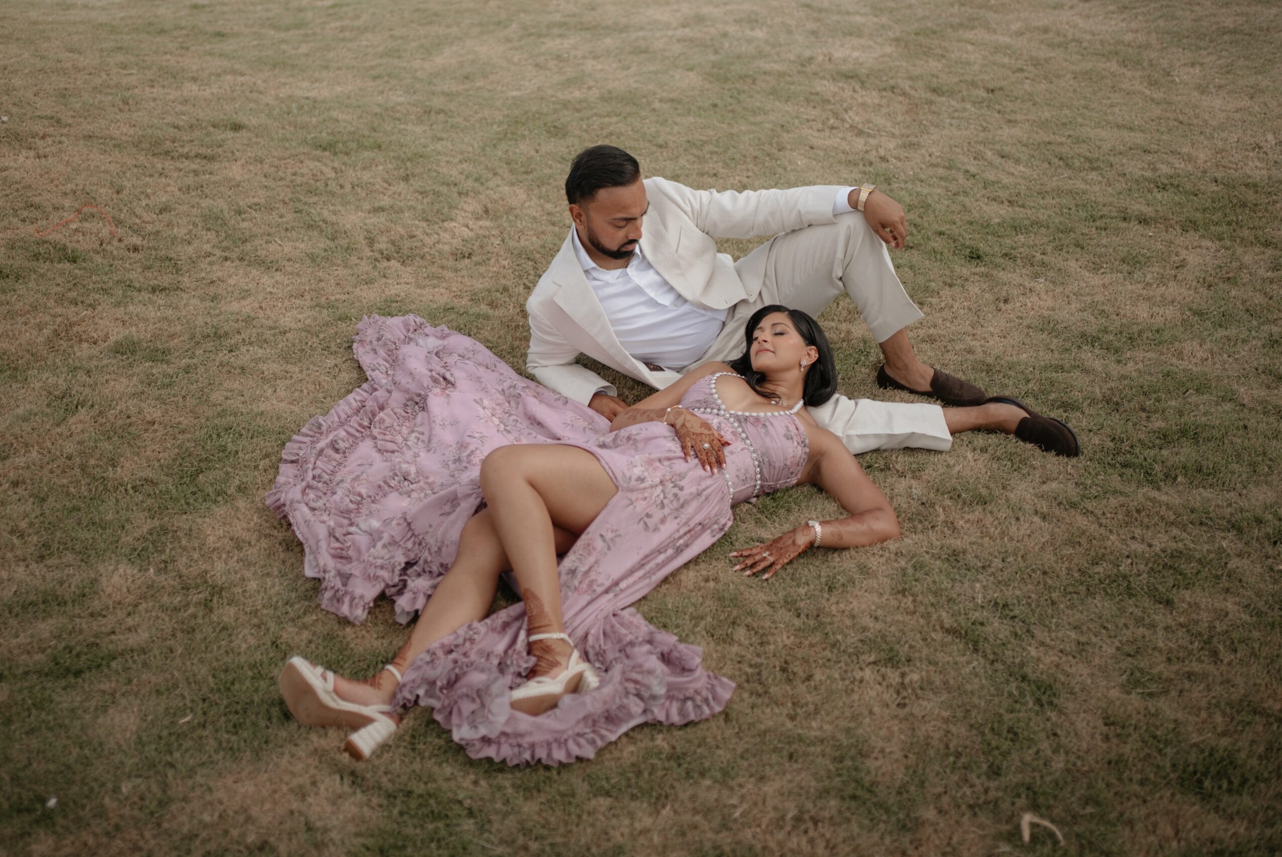 A man and woman lie on grass, the woman in a pink dress, the man in a white suit, holding hands.