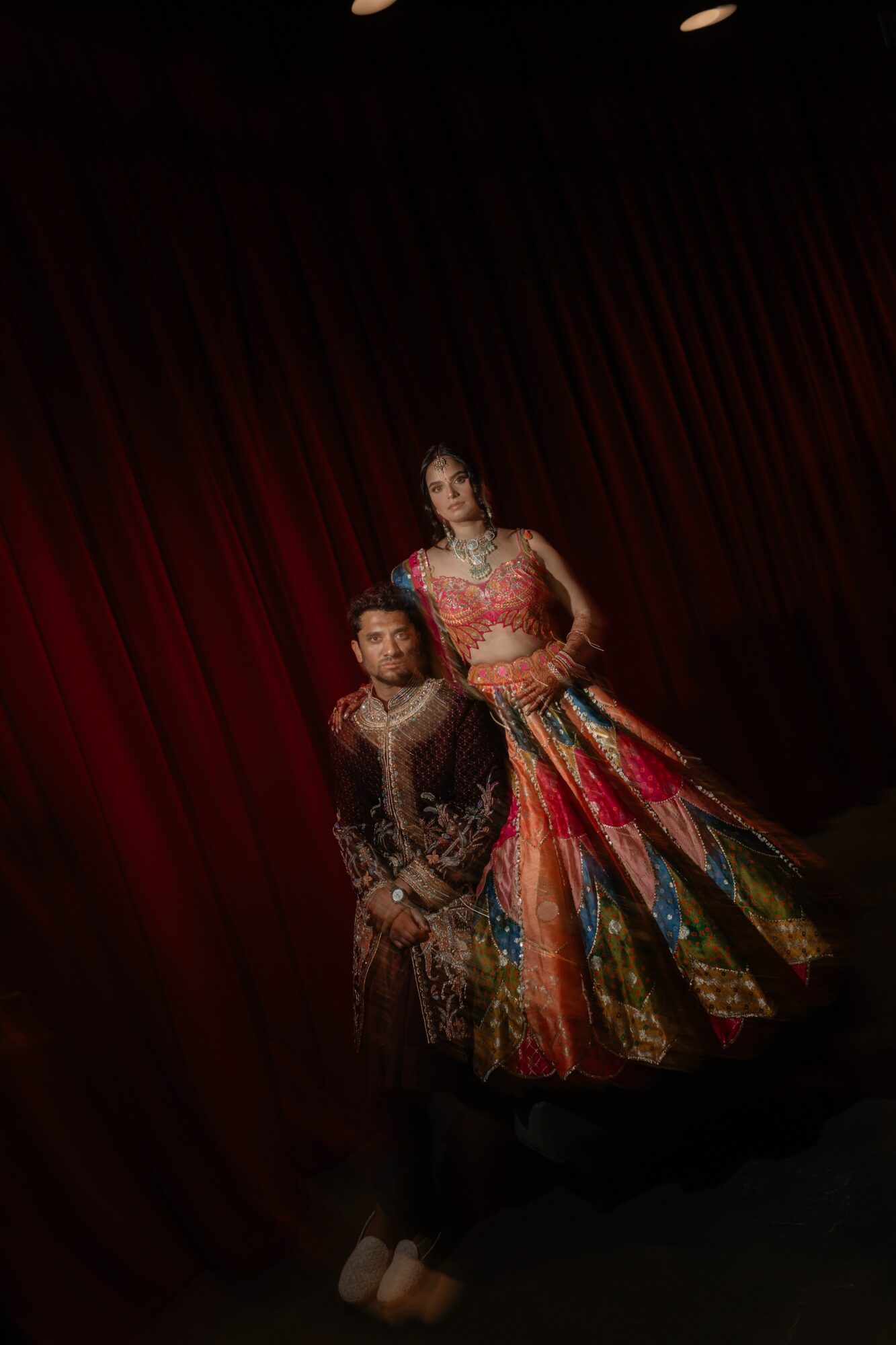 Two performers in colorful costumes stand on stage with a red curtain background.