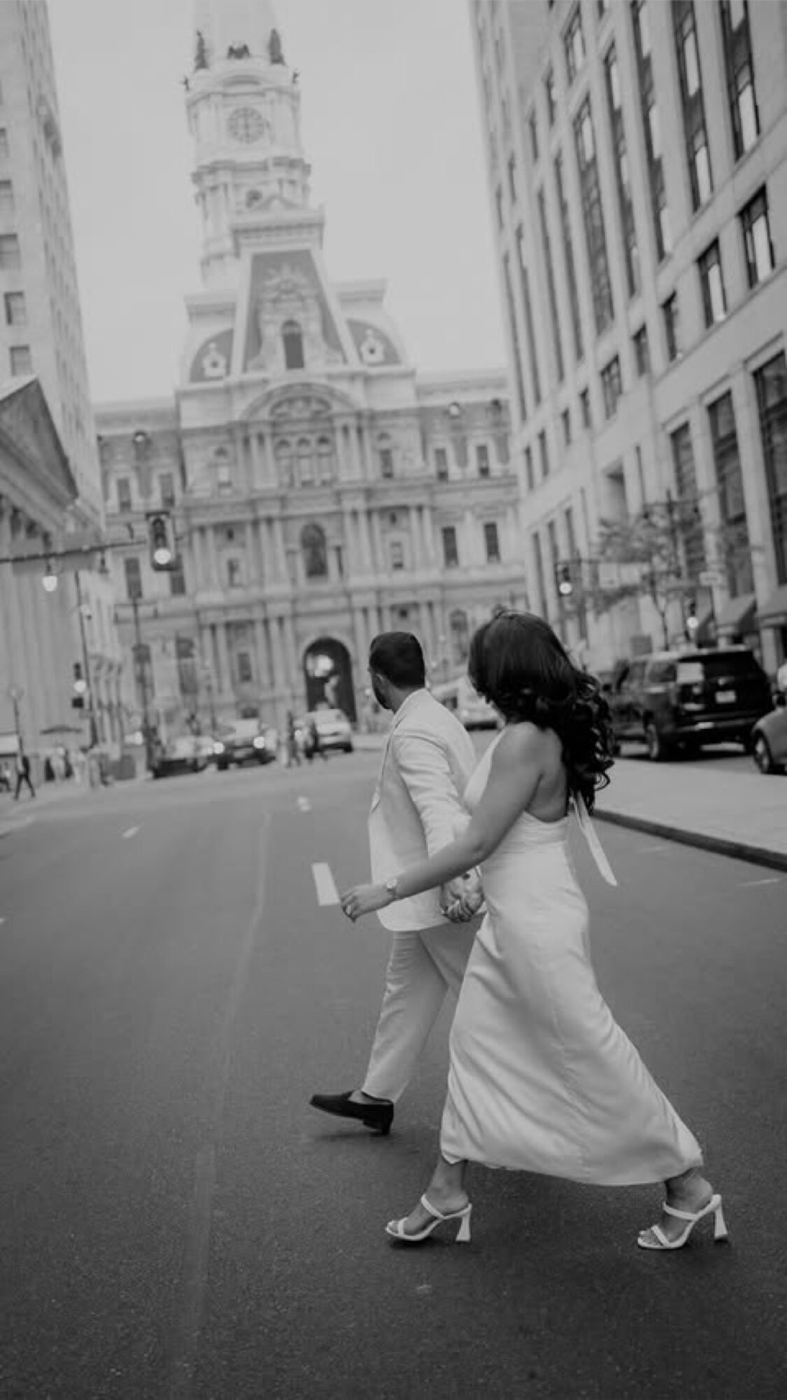 A couple crossing a city street with historic building in background, woman in white dress, man in light suit, black and white photo.