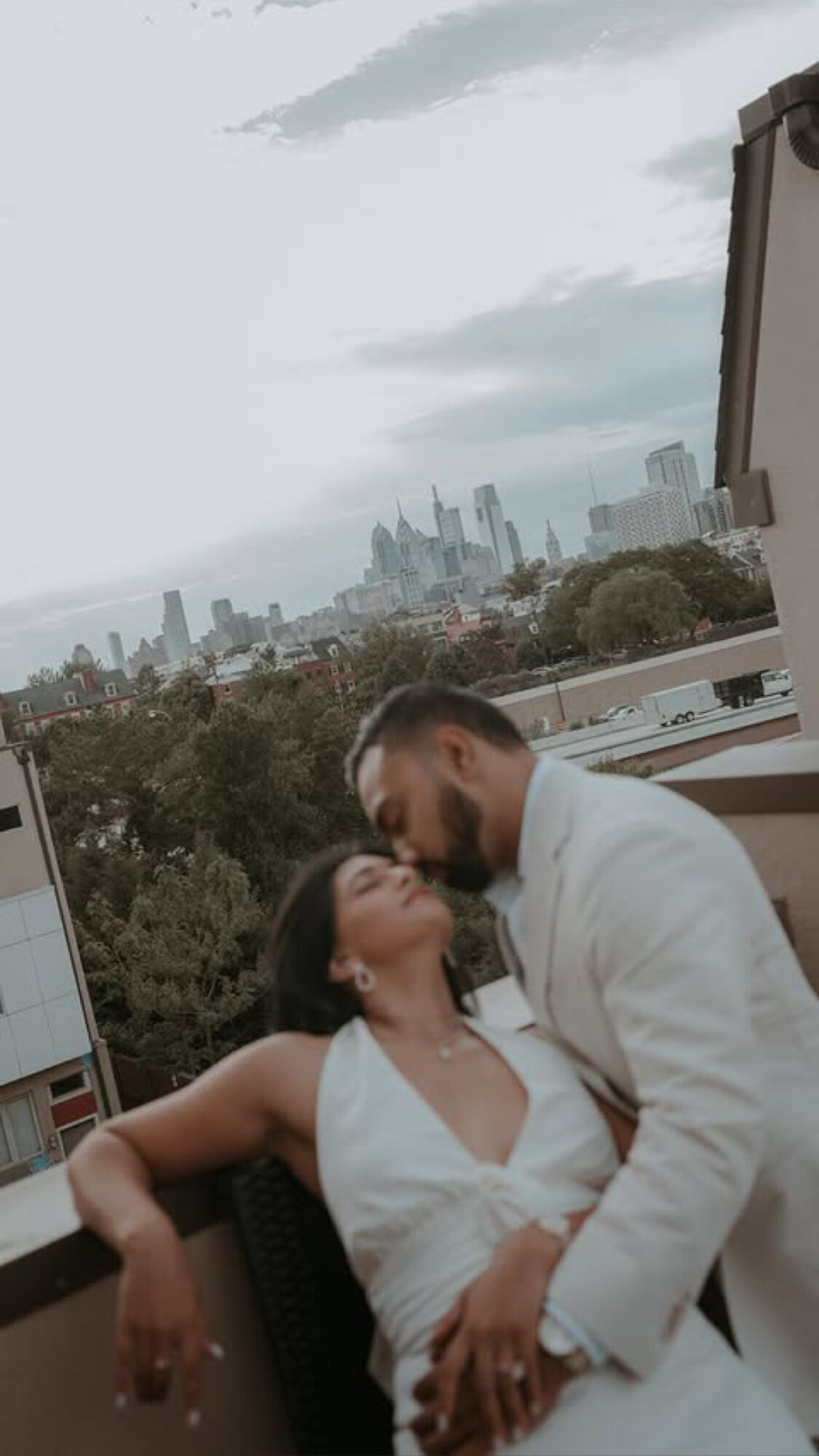 Two people embrace on a balcony with a city skyline in the background, overcast sky, one in white dress, one in white suit.