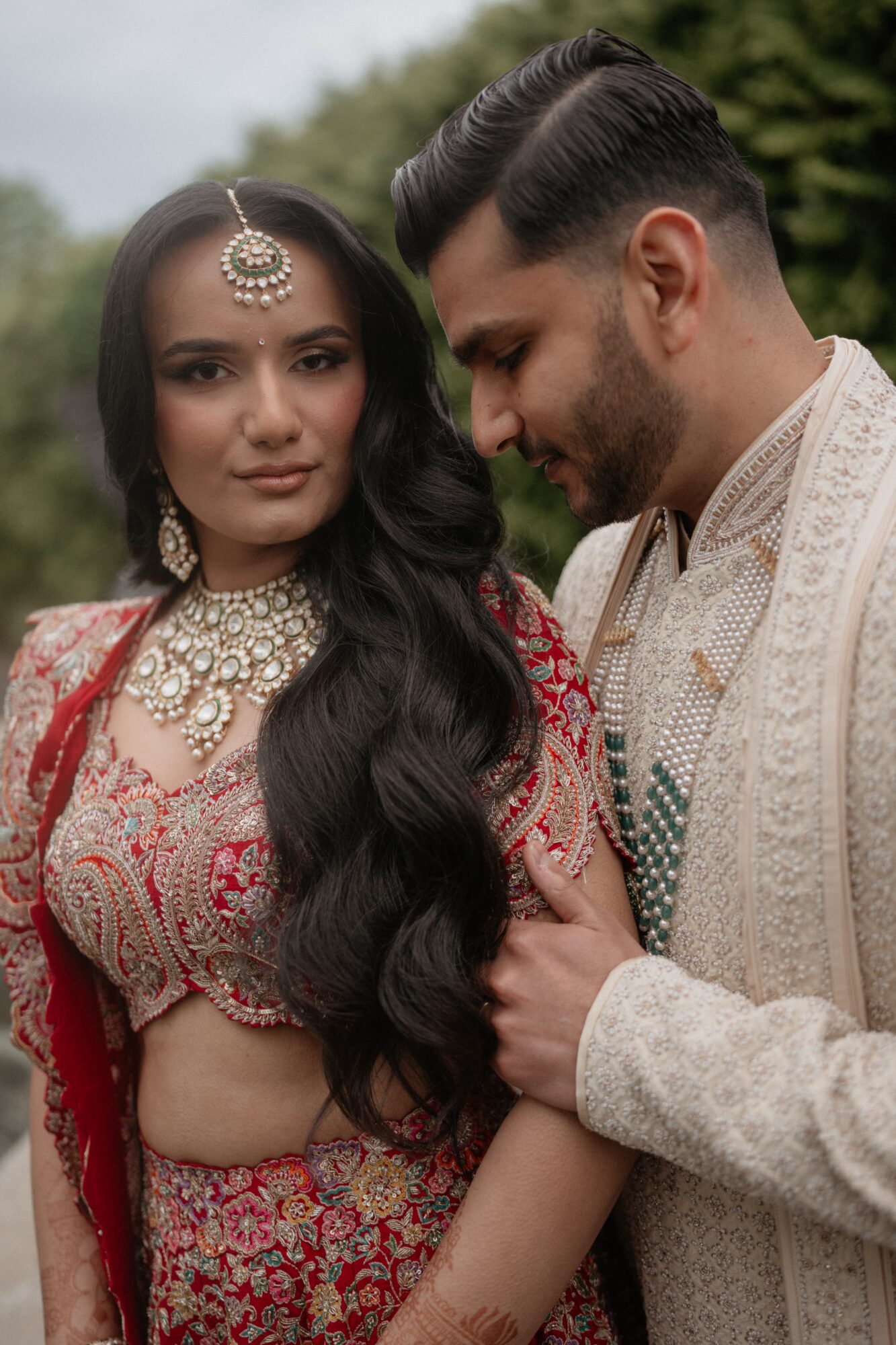 A couple dressed in traditional attire outdoors, with the woman wearing jewelry and the man looking at her affectionately.