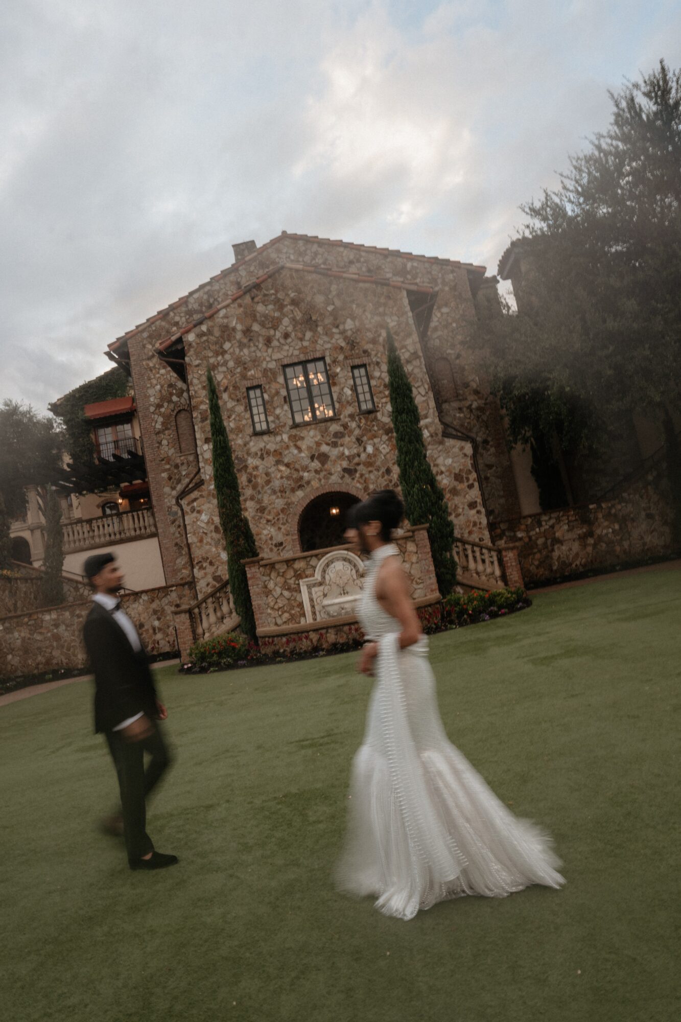 Two people in wedding attire standing on grass in front of a stone building with windows and trees.