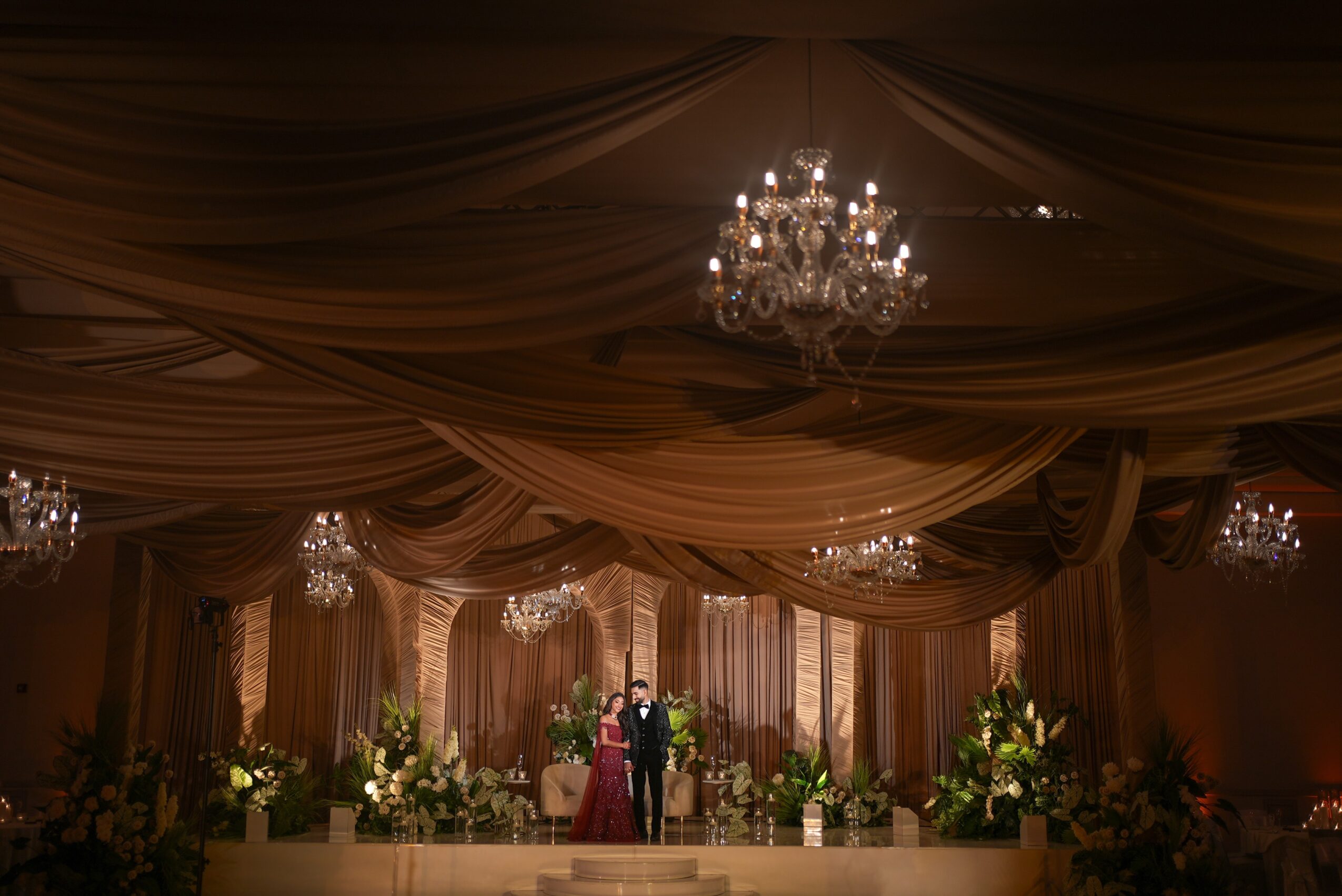 People dancing in a decorated ballroom with chandeliers, draped ceiling, and floral arrangements, illuminated warmly.