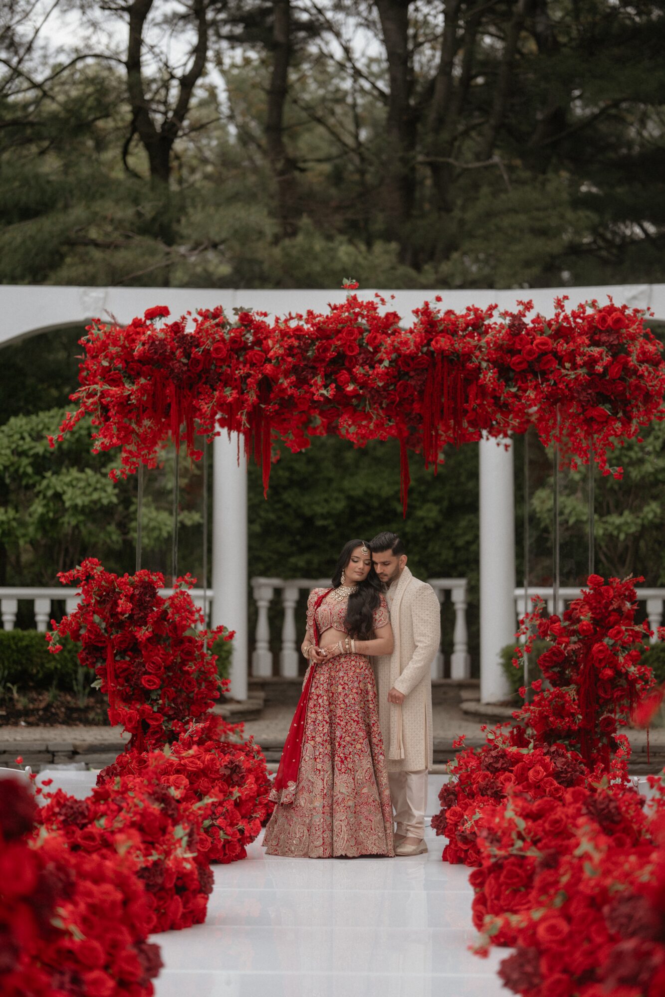 Couple standing under a decorated archway with red flowers, surrounded by red floral arrangements, outdoors with trees in background.
